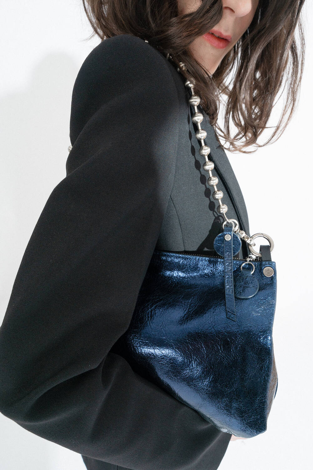 A person in a black blazer elegantly holds an INA KENT handbag—a small, shiny blue accessory with a silver chain strap.