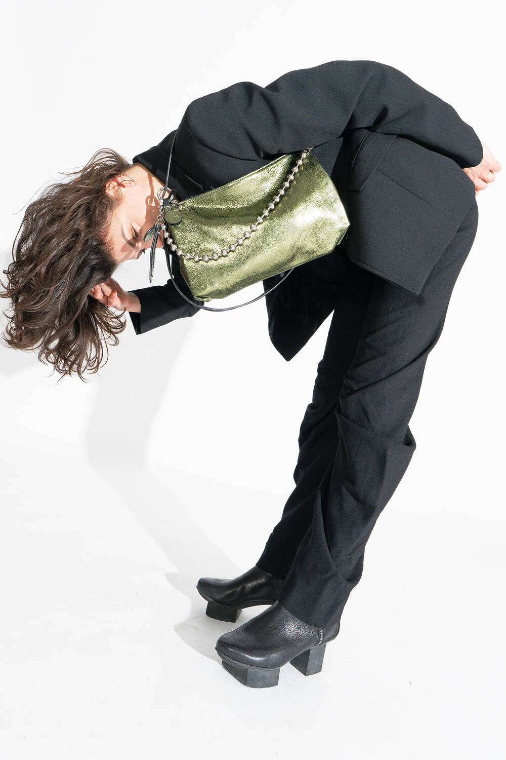 Eine Person in einem schwarzen Anzug und Plateaustiefeln beugt sich nach vorne und präsentiert eine grüne Handtasche mit Kettenriemen von INA KENT.