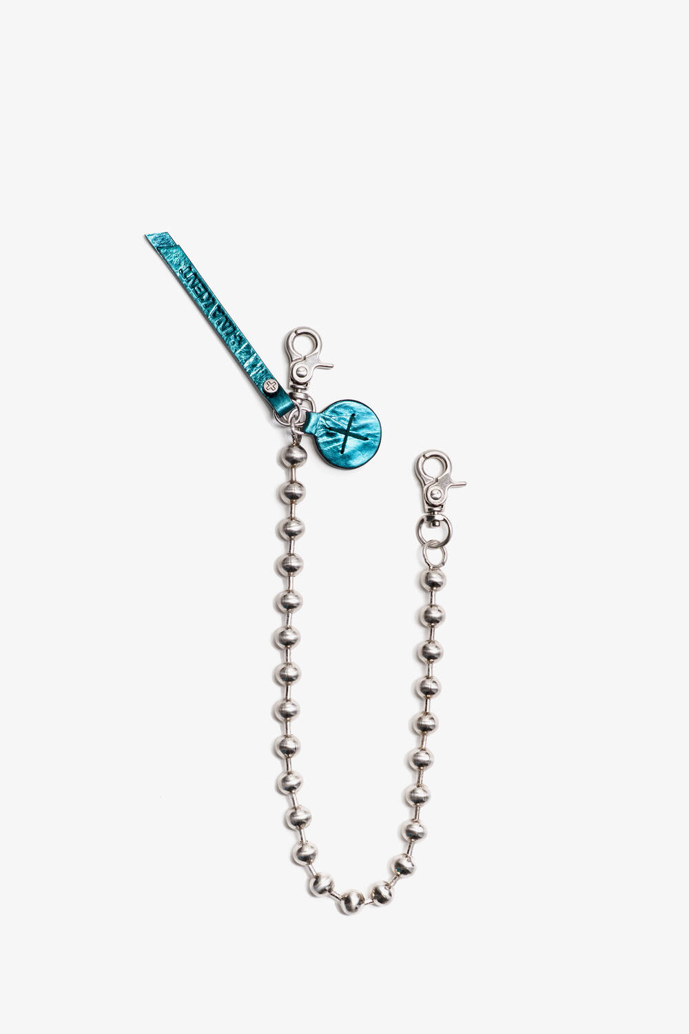 Metallisches Kugelkettenarmband mit Karabinerverschluss, einem blauen Etikett und einem runden Anhänger mit eingraviertem INA KENT-Symbol.