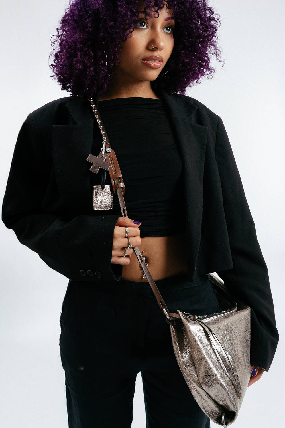 Eine Frau mit lockigem lila Haar trägt einen schwarzen kurzen Blazer und ein schwarzes Top, eine schwarze Hose und eine metallische Umhängetasche von INA KENT an einem breiten Riemen.