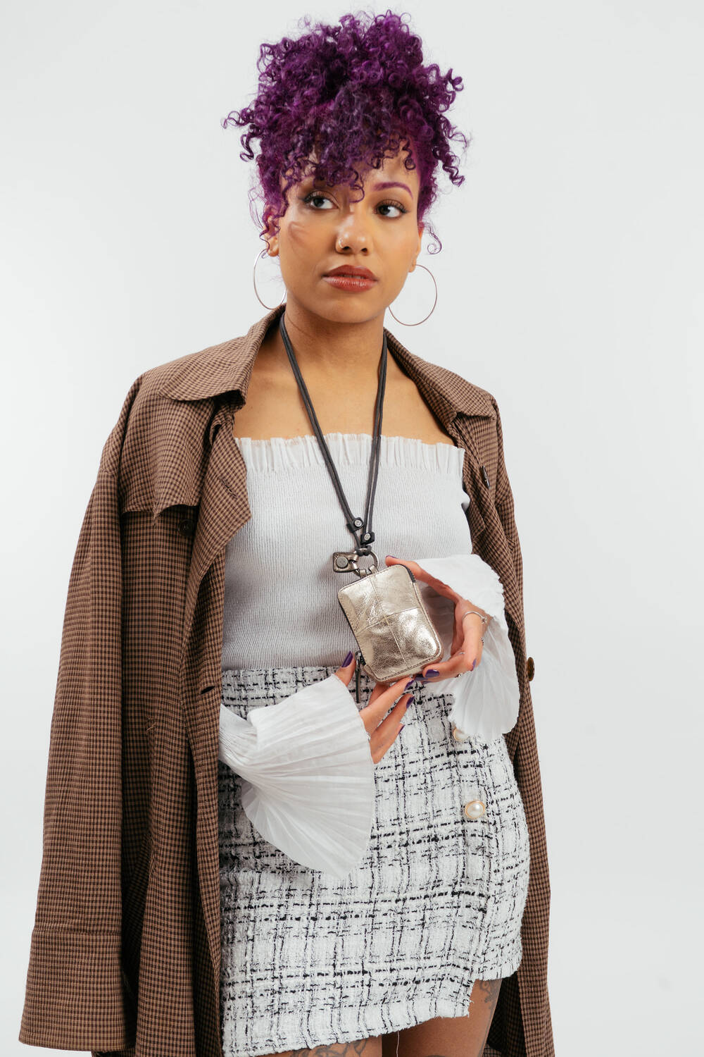 Eine Frau mit lockigem lila Haar trägt einen braunen karierten Mantel über einem weißen Oberteil und einem Tweedrock und hält eine kleine silberne Tasche von INA KENT an einem schwarzen Riemen um den Hals.