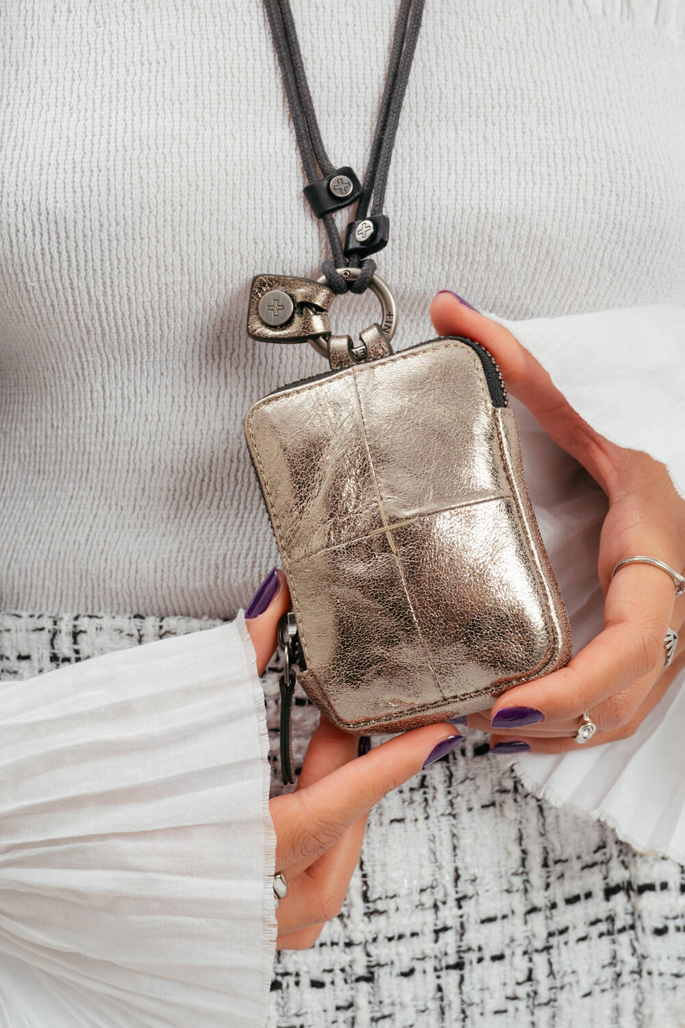Eine Person hält einen kleinen metallisch-goldenen INA KENT-Beutel mit Reißverschluss, der an einem schwarzen Band um den Hals befestigt ist. Sie hat manikürte Nägel und trägt eine strukturierte weiße Bluse.