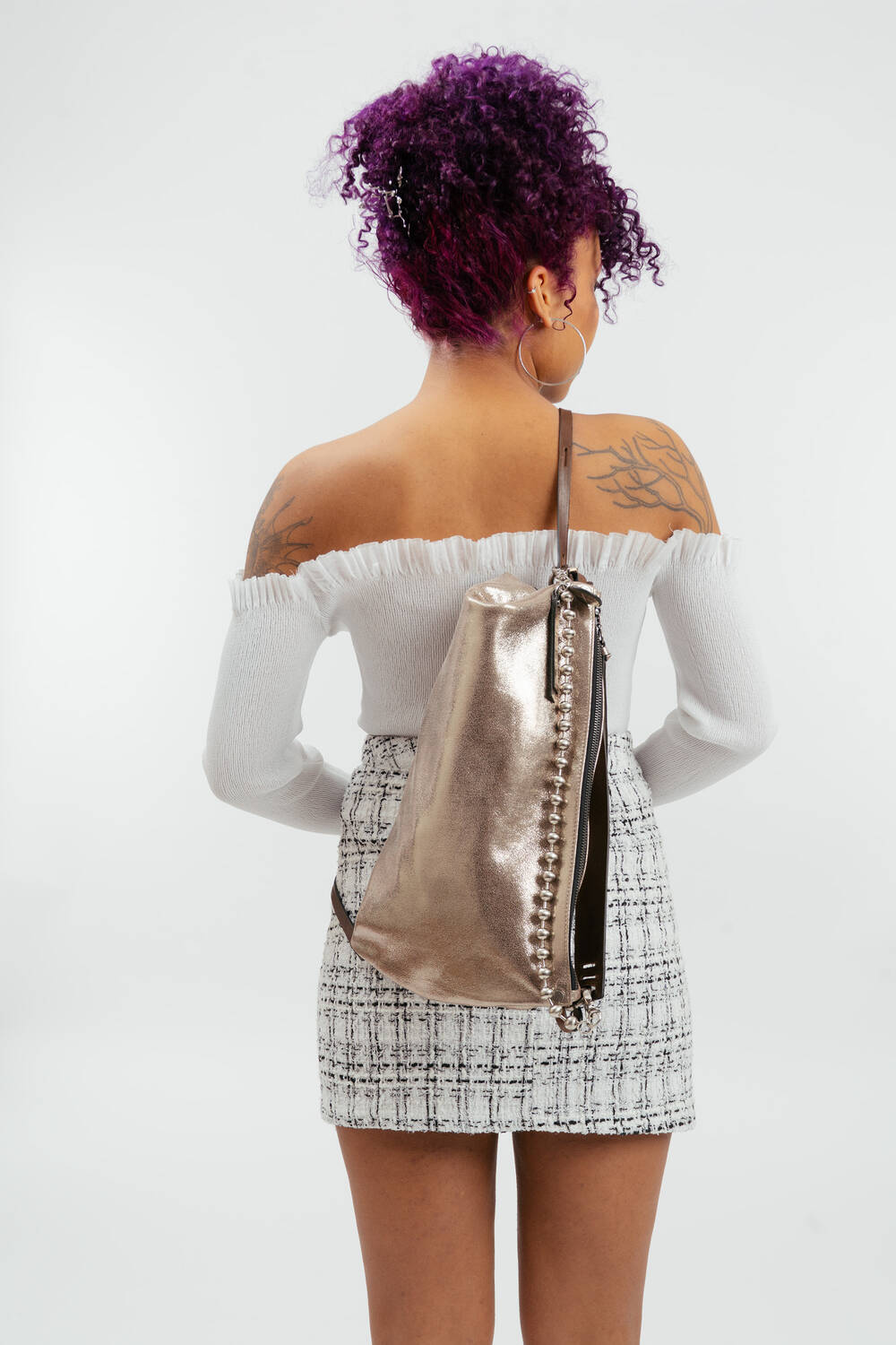 Eine Person mit lockigem lila Haar, die ein schulterfreies weißes Oberteil und einen karierten Rock trägt, steht mit dem Rücken zur Wand und trägt einen metallischen INA KENT-Rucksack.