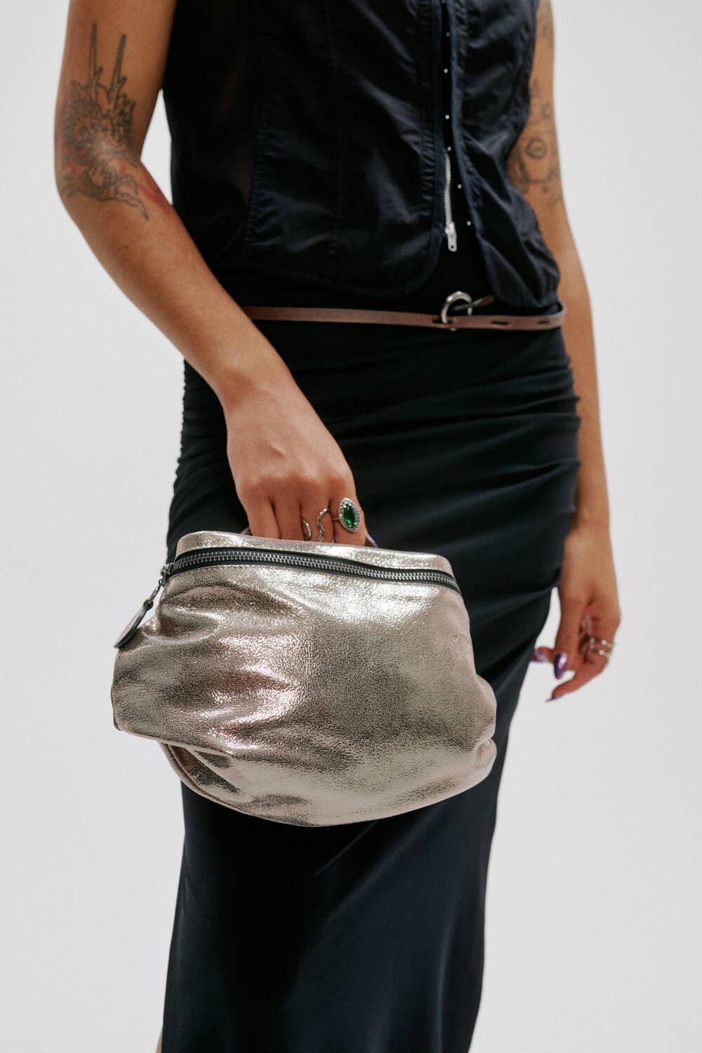 Eine Person in einem ärmellosen schwarzen Top und Rock hält eine metallisch glänzende silberne Clutch von INA KENT mit Reißverschluss; auf ihrer Hand und ihrem Arm sind Tattoos und Schmuck zu sehen.