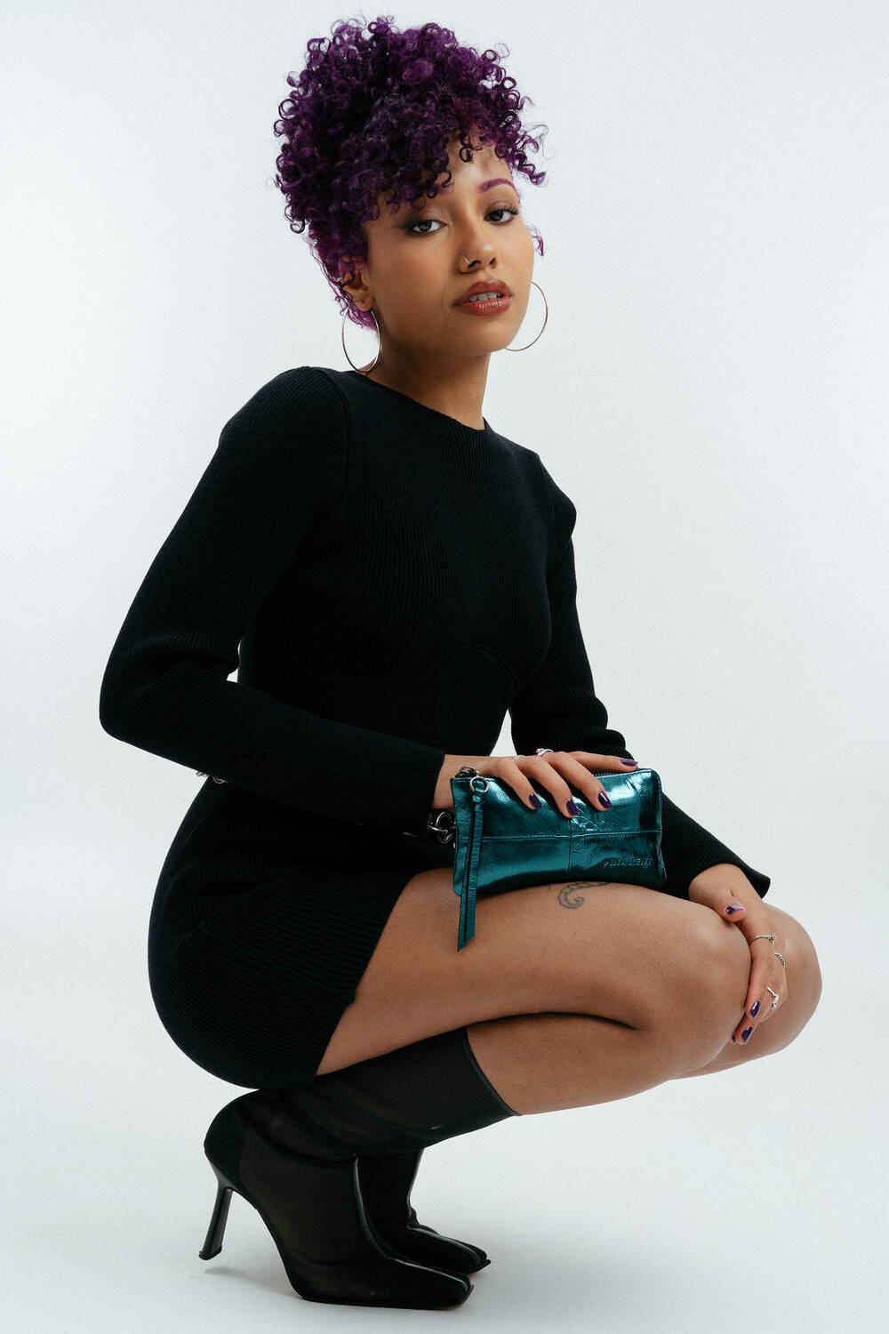 Eine Frau mit lockigem lila Haar, die ein schwarzes Langarmkleid und schwarze High Heels trägt, hockt und hält vor einem schlichten weißen Hintergrund eine kleine metallisch-blaugrüne Handtasche von INA KENT.