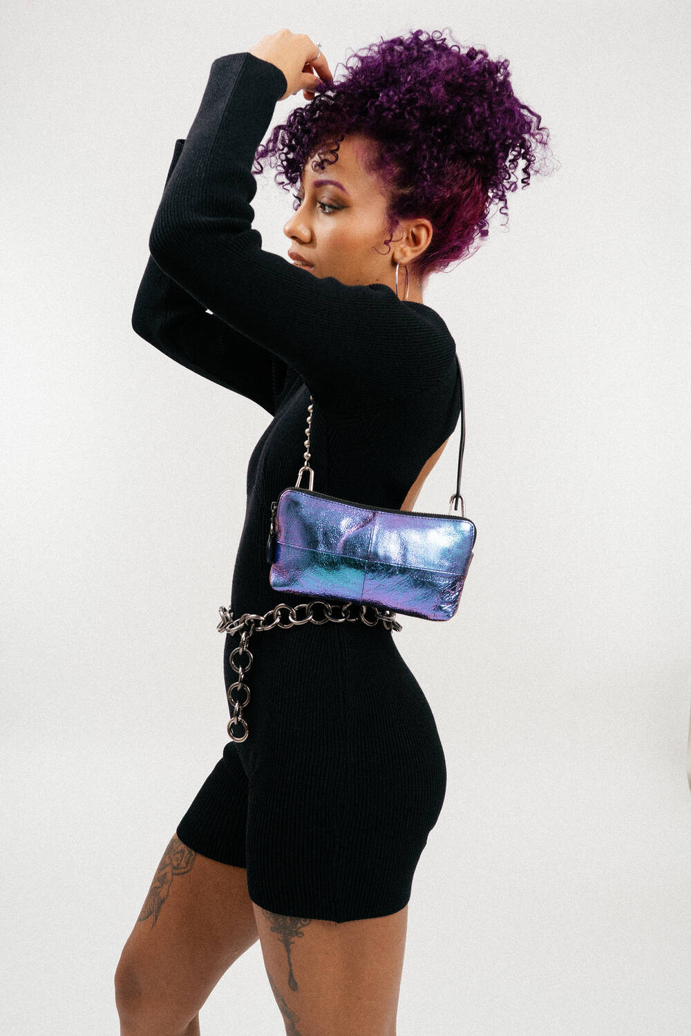 A person with curly purple hair wears a black long-sleeve outfit and carries an INA KENT metallic purple purse with a chain strap around their waist.