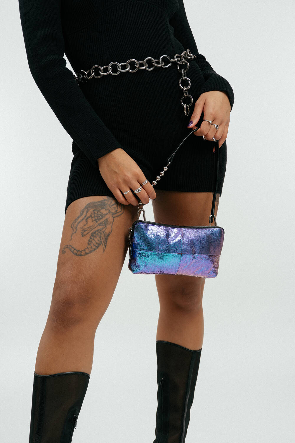 Person wearing a black dress, chain belt, and black boots holds an iridescent INA KENT purse; a snake tattoo is visible on their thigh.
