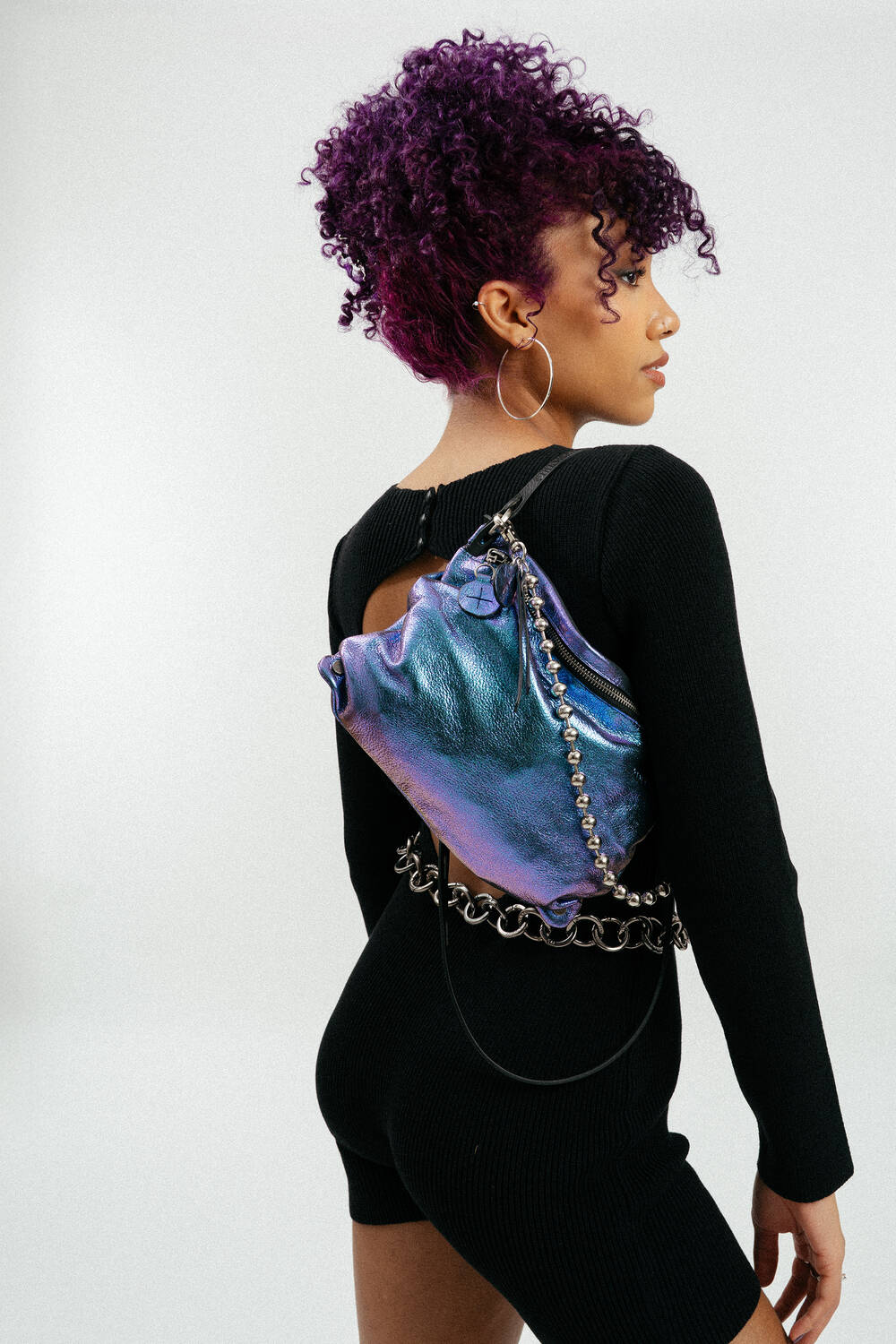 A person with curly purple hair and hoop earrings wears a black outfit and carries an INA KENT metallic purple bag with a chain strap over their back.
