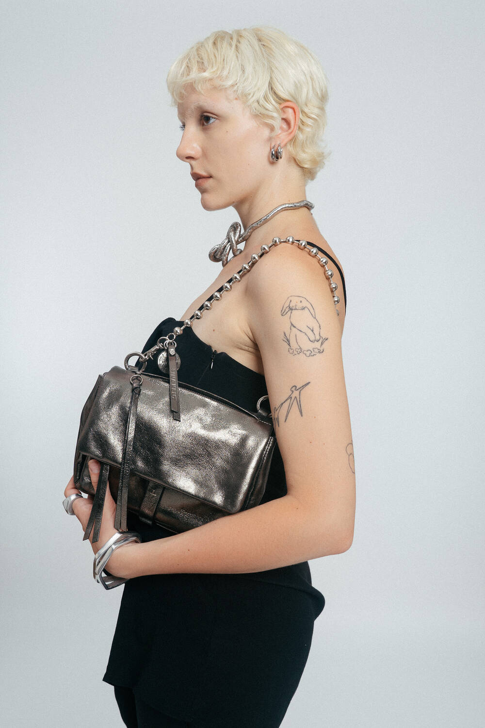 Person with short blond hair, silver jewelry, black outfit, and visible tattoos on their arm holds an INA KENT metallic handbag with a chain strap against a plain background.