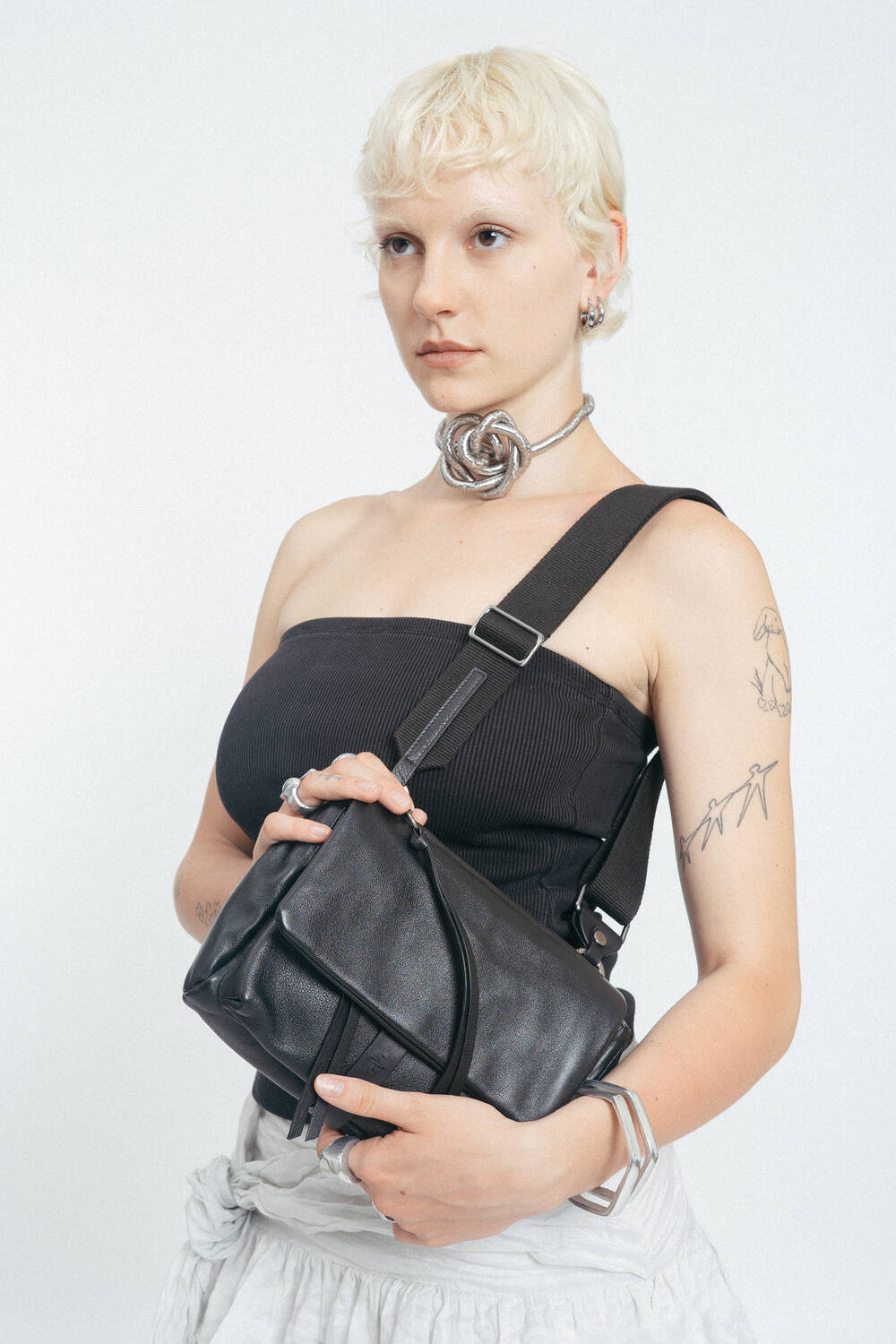 Eine Person mit kurzen blonden Haaren trägt ein schwarzes Schlauchtop, silberne Accessoires und hält eine schwarze INA KENT-Umhängetasche vor einem schlichten weißen Hintergrund.
