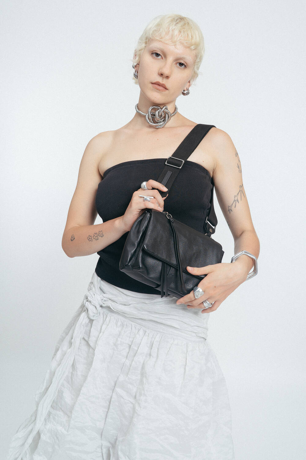 Person mit kurzen blonden Haaren trägt ein schwarzes trägerloses Oberteil, einen weißen Rock, klobigen Schmuck und trägt eine schwarze INA KENT Crossbody Bag vor einem einfarbigen Hintergrund.