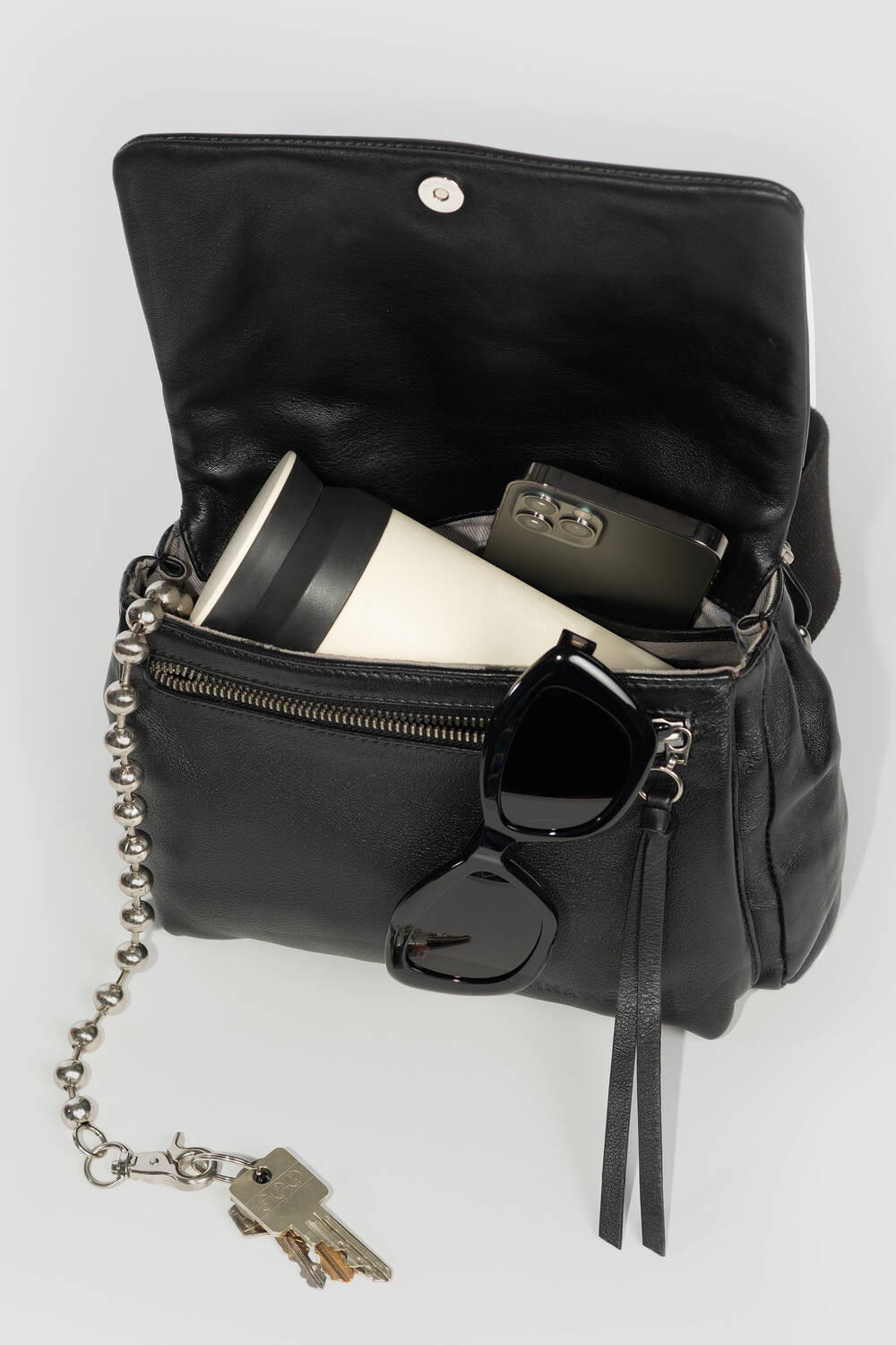 Ein schwarzes Portemonnaie von INA KENT mit einem Reißverschlussfach enthält einen Reisebecher und ein Smartphone. Die Sonnenbrille hängt am Reißverschluss, und ein Schlüsselbund mit Schlüsseln ist an der Tasche befestigt.
