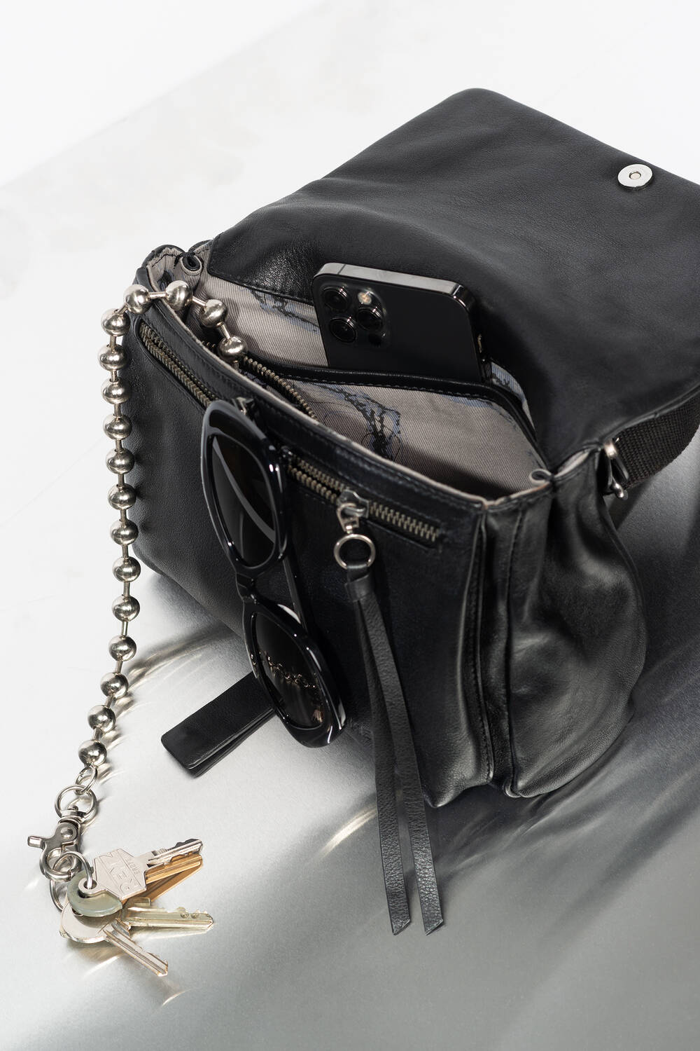 Eine schwarze Ledertasche von INA KENT liegt offen auf einer hellen Unterlage und enthält ein Telefon, eine Sonnenbrille und eine Kette mit Schlüsseln daran.