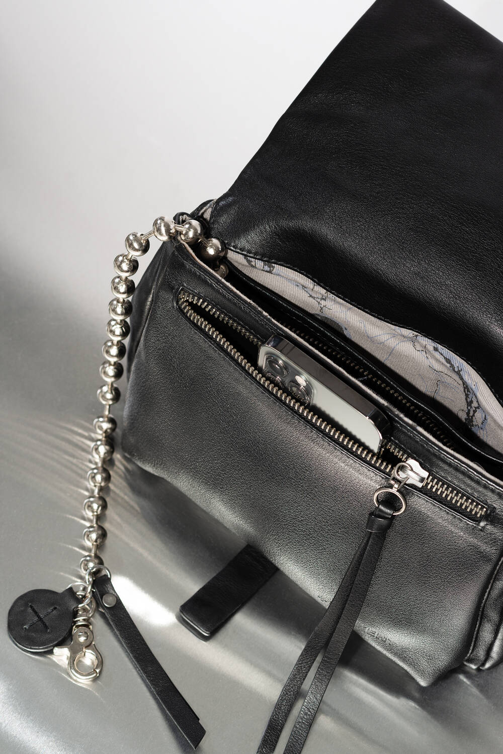 INA KENT Tasche aus schwarzem Leder mit metallischer Kugelkette, Reißverschluss und einer offenen Tasche, in der ein silbernes Smartphone steckt.
