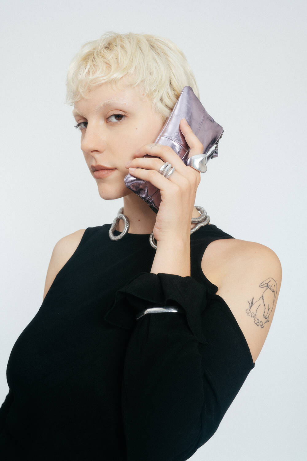 Frau mit kurzen blonden Haaren hält eine lilafarbene Metalltasche von INA KENT an ihr Ohr. Sie trägt ein schwarzes Oberteil mit ausgeschnittenen Schultern und Silberschmuck und zeigt ein Kaninchentattoo auf ihrem Oberarm.