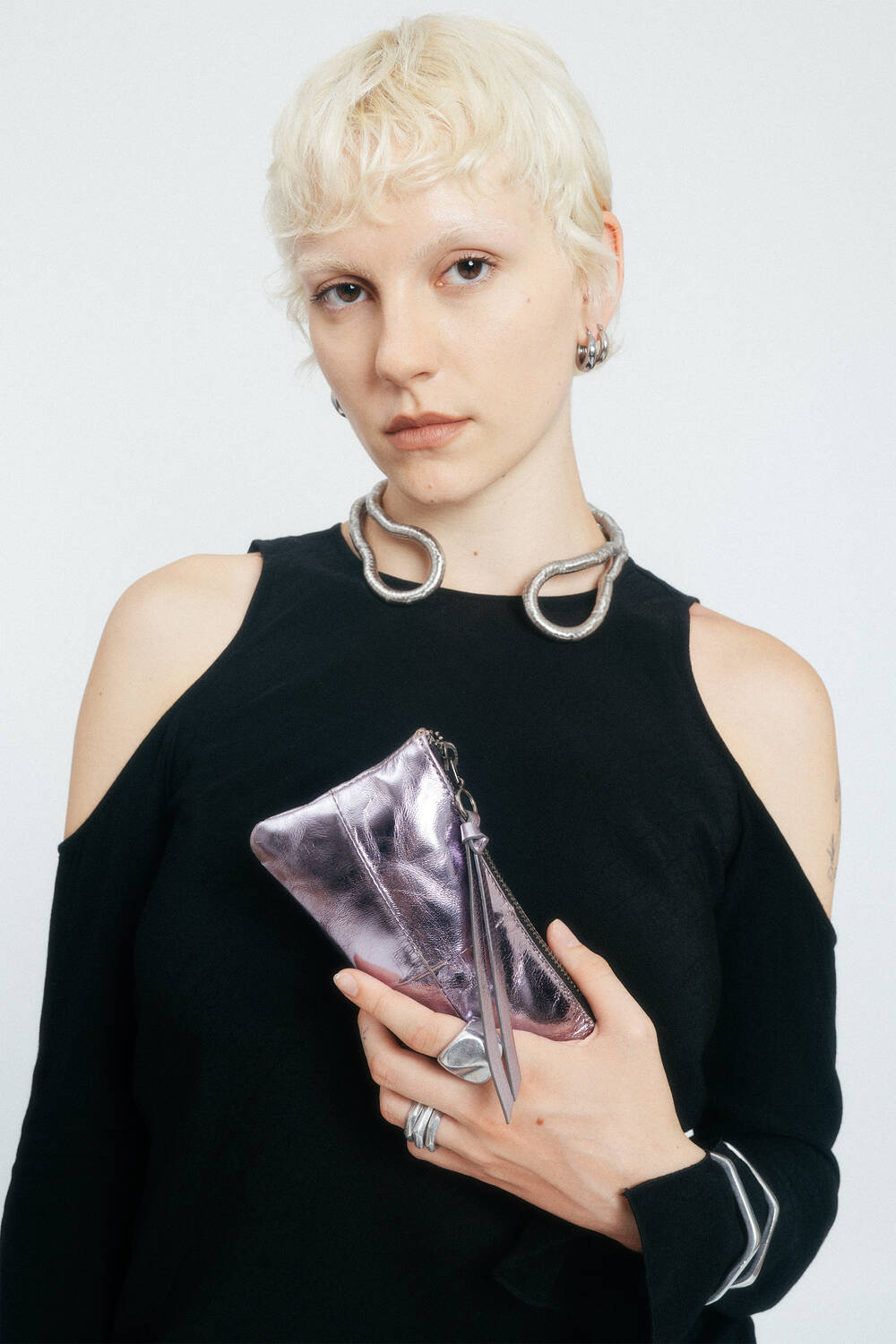 Person mit kurzen blonden Haaren, die ein schwarzes Oberteil trägt, hält eine lilafarbene INA KENT-Tasche in der Hand; dazu trägt sie klobigen Schmuck, darunter Reifenohrringe und eine große silberne Halskette.