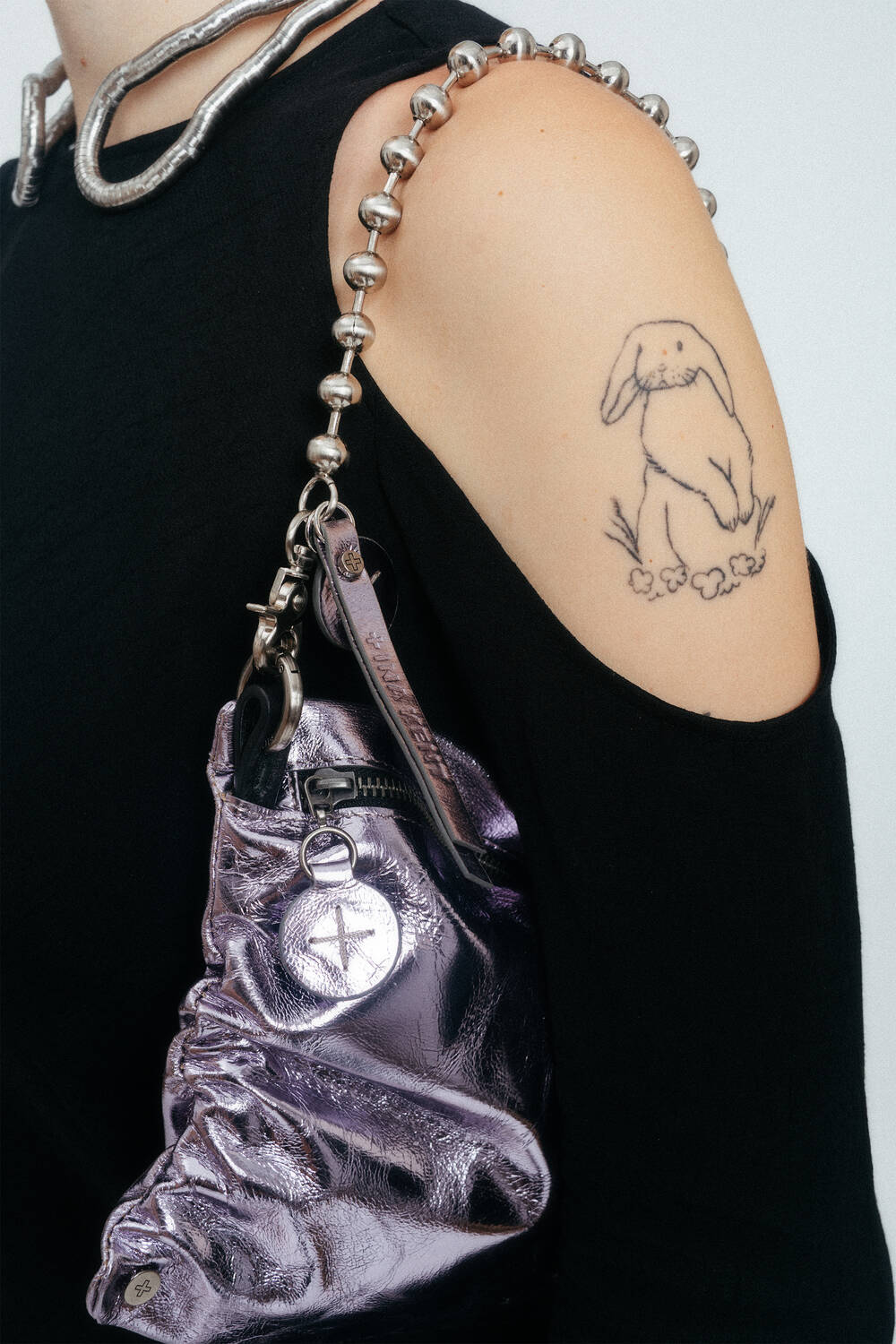 Person wearing a black top with cut-out shoulders, a bunny tattoo on upper arm, and carrying an INA KENT metallic purple bag with silver chain strap.