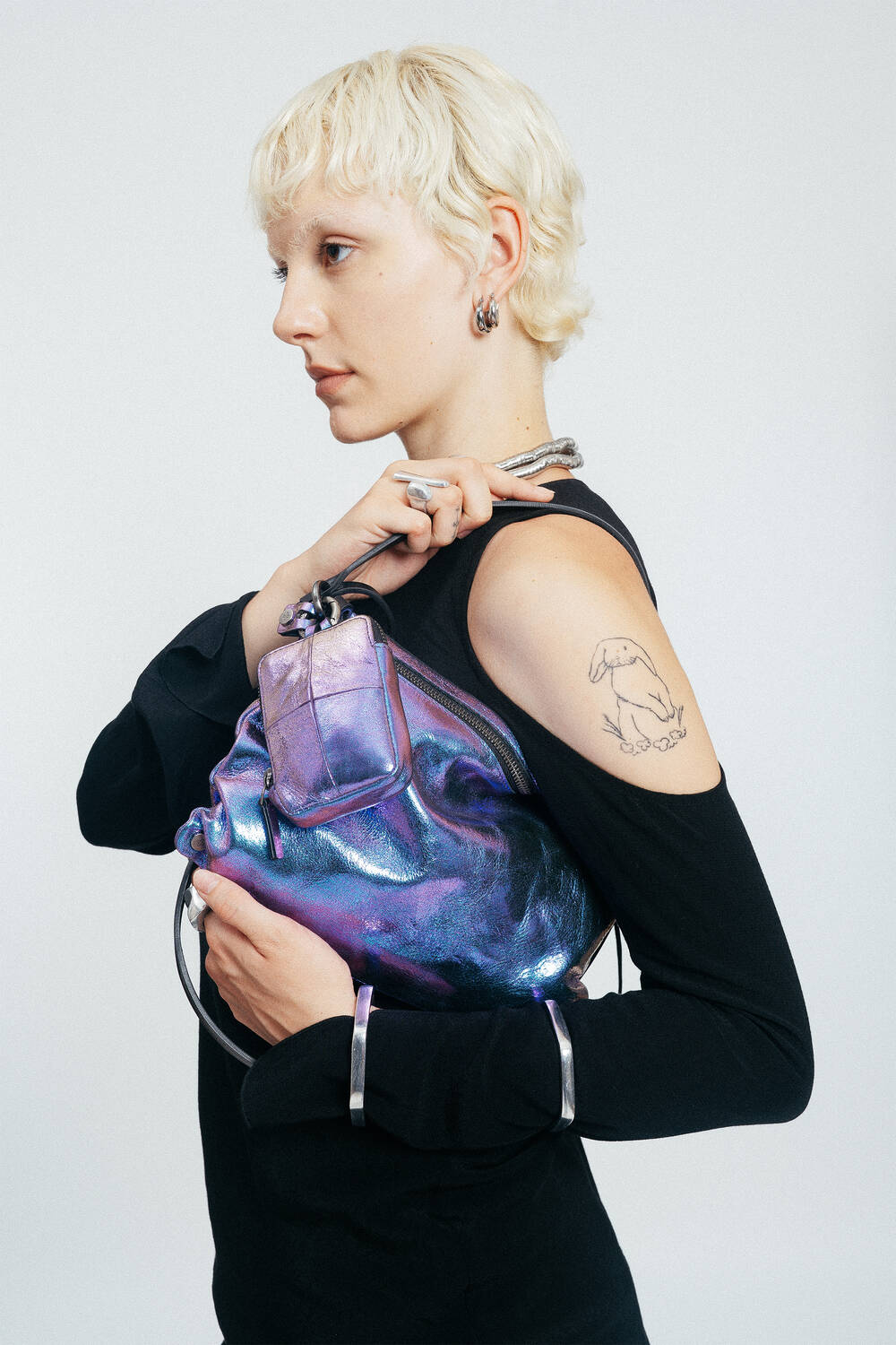 Person with short blonde hair holds an iridescent purple INA KENT handbag, displaying a rabbit tattoo on their upper arm. They wear a black long-sleeve top and silver jewelry.