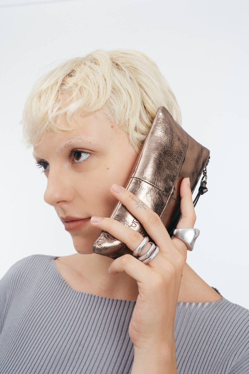 Person mit kurzen blonden Haaren, die eine INA KENT-Metalltasche wie ein Telefon an ihr Ohr hält, mit einem gerippten grauen Oberteil und silbernen Ringen, vor einem schlichten hellen Hintergrund.