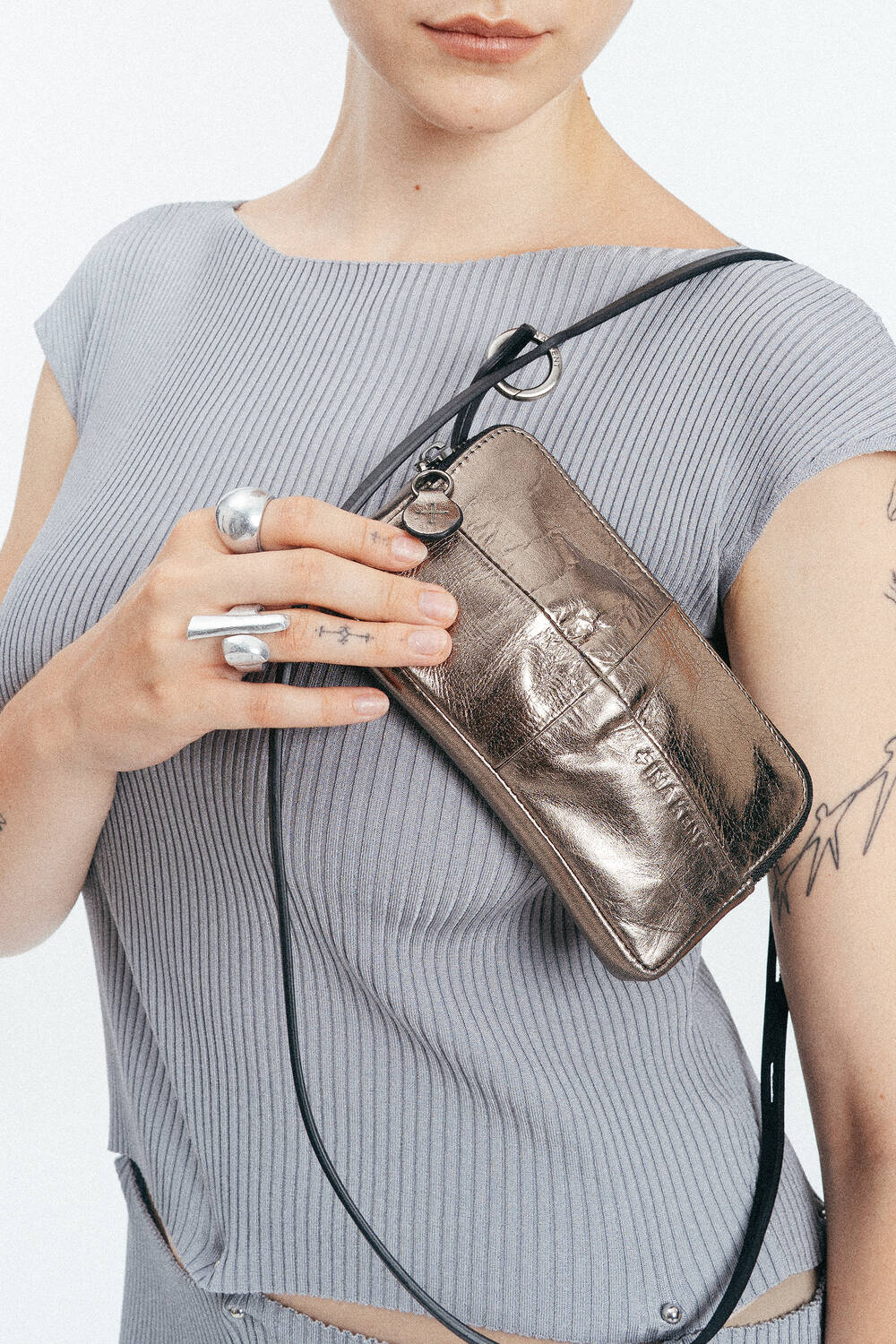 Die Person, die ein geripptes graues Oberteil trägt, hält eine INA KENT-Metallic-Crossbody-Tasche dicht vor der Brust und zeigt silberne Ringe und sichtbare Tätowierungen auf ihrer Hand und ihrem Arm.