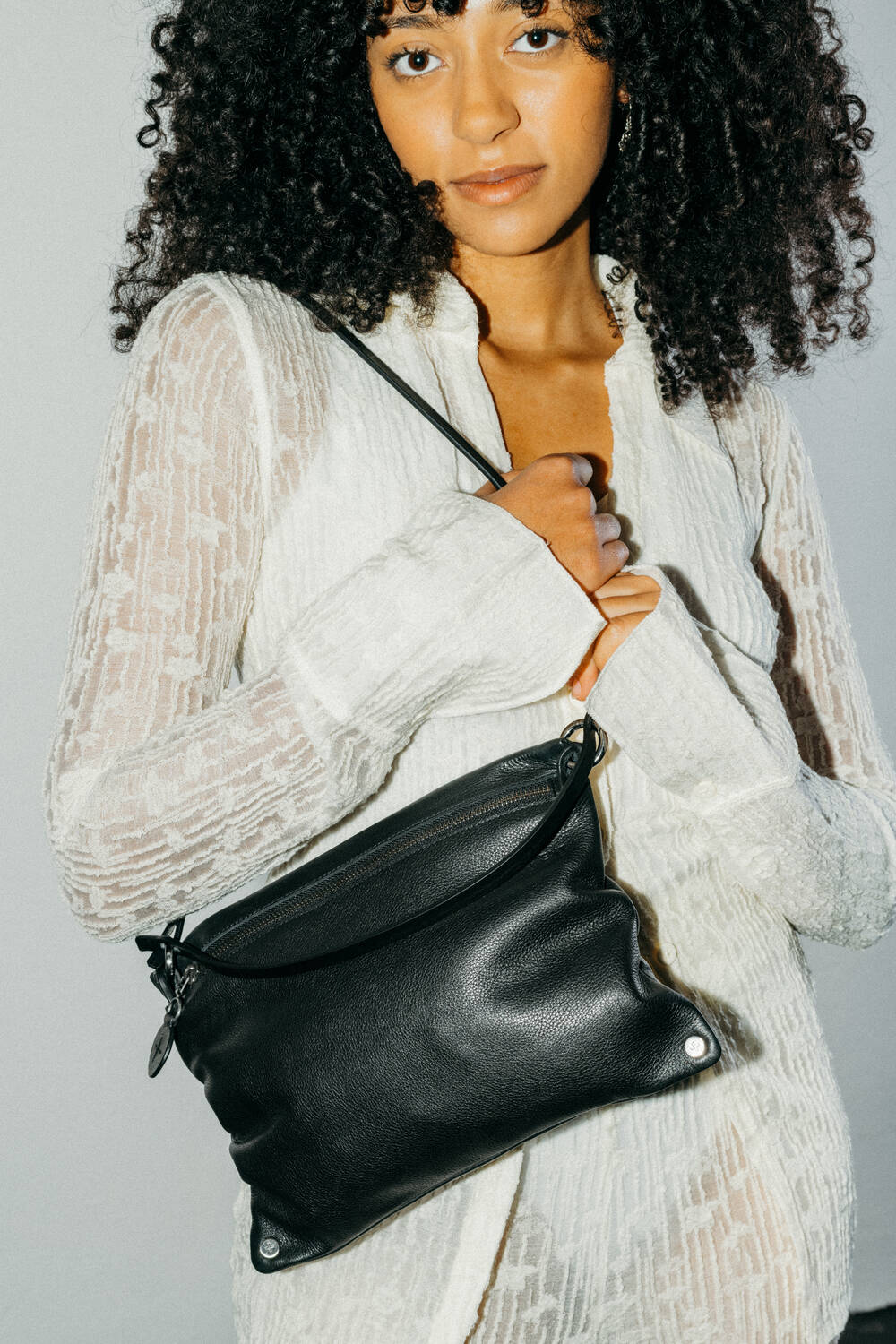 A woman wearing a sheer white dress holds an INA KENT black leather crossbody bag with metal accents, standing against a plain light background.