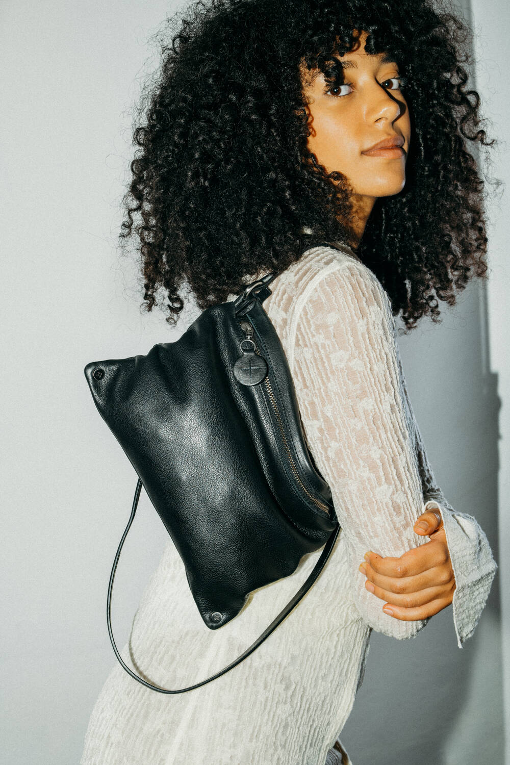 A person with curly hair wearing a white textured dress and carrying an INA KENT black leather backpack over one shoulder, looking towards the camera.