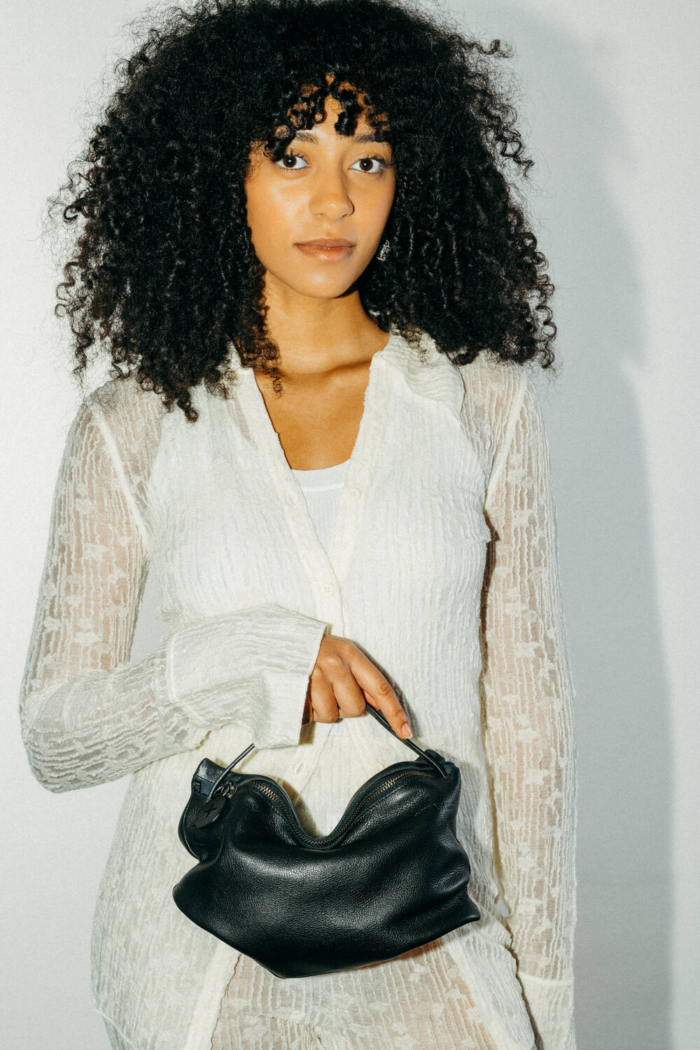 A woman with curly hair wearing a sheer white long-sleeve top holds an INA KENT black leather handbag in front of her against a plain white background.