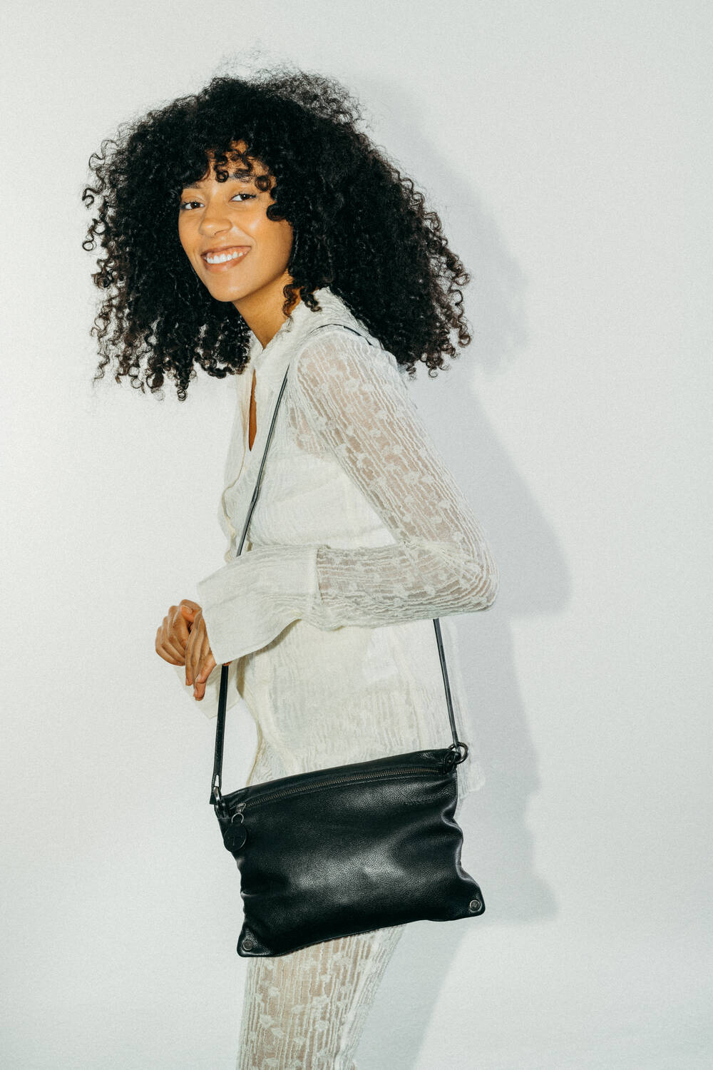 A woman with curly hair wearing a textured white outfit holds an INA KENT black crossbody bag and smiles, standing against a plain light background.