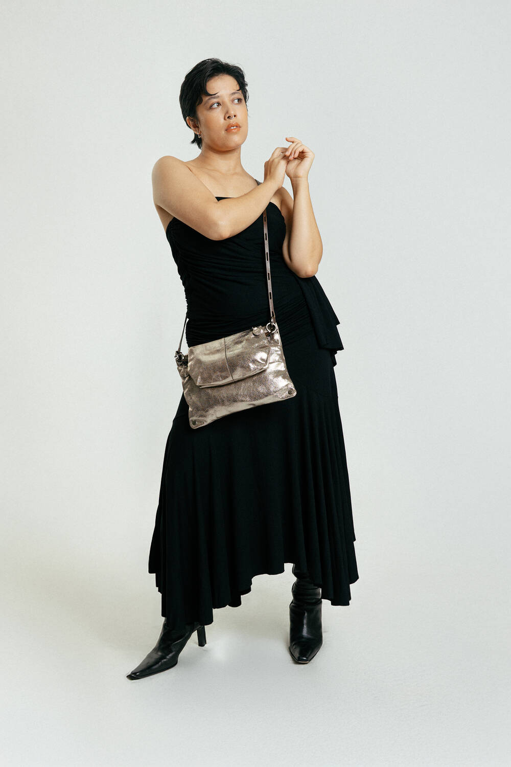 Person, die ein schwarzes One-Shoulder-Kleid und schwarze Stiefel trägt und eine INA KENT Metallic-Crossbody-Tasche in der Hand hält, steht vor einem schlichten weißen Hintergrund.