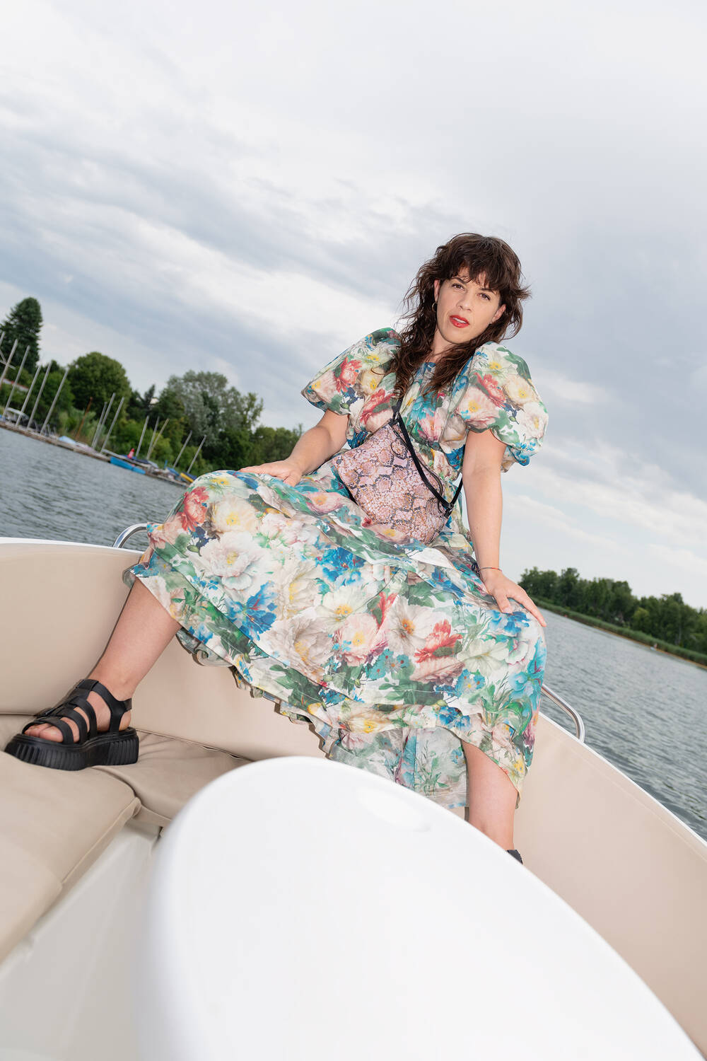 Eine Frau in einem geblümten Kleid und schwarzen Sandalen, die ihre modische INA KENT-Tasche trägt, sitzt am Bug eines Bootes mit einem See und Bäumen im Hintergrund unter einem bewölkten Himmel.