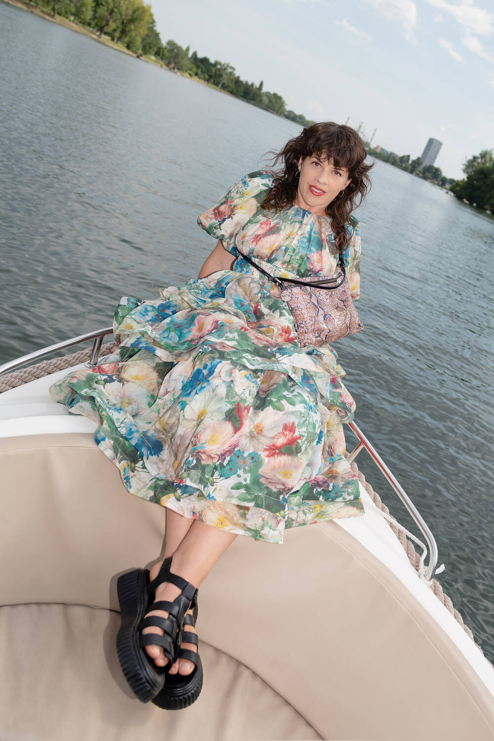 Eine Frau in einem geblümten Kleid und schwarzen Sandalen, die eine INA KENT-Tasche trägt, sitzt auf dem Rand eines Bootes mit einer Wasserfläche und Bäumen im Hintergrund.