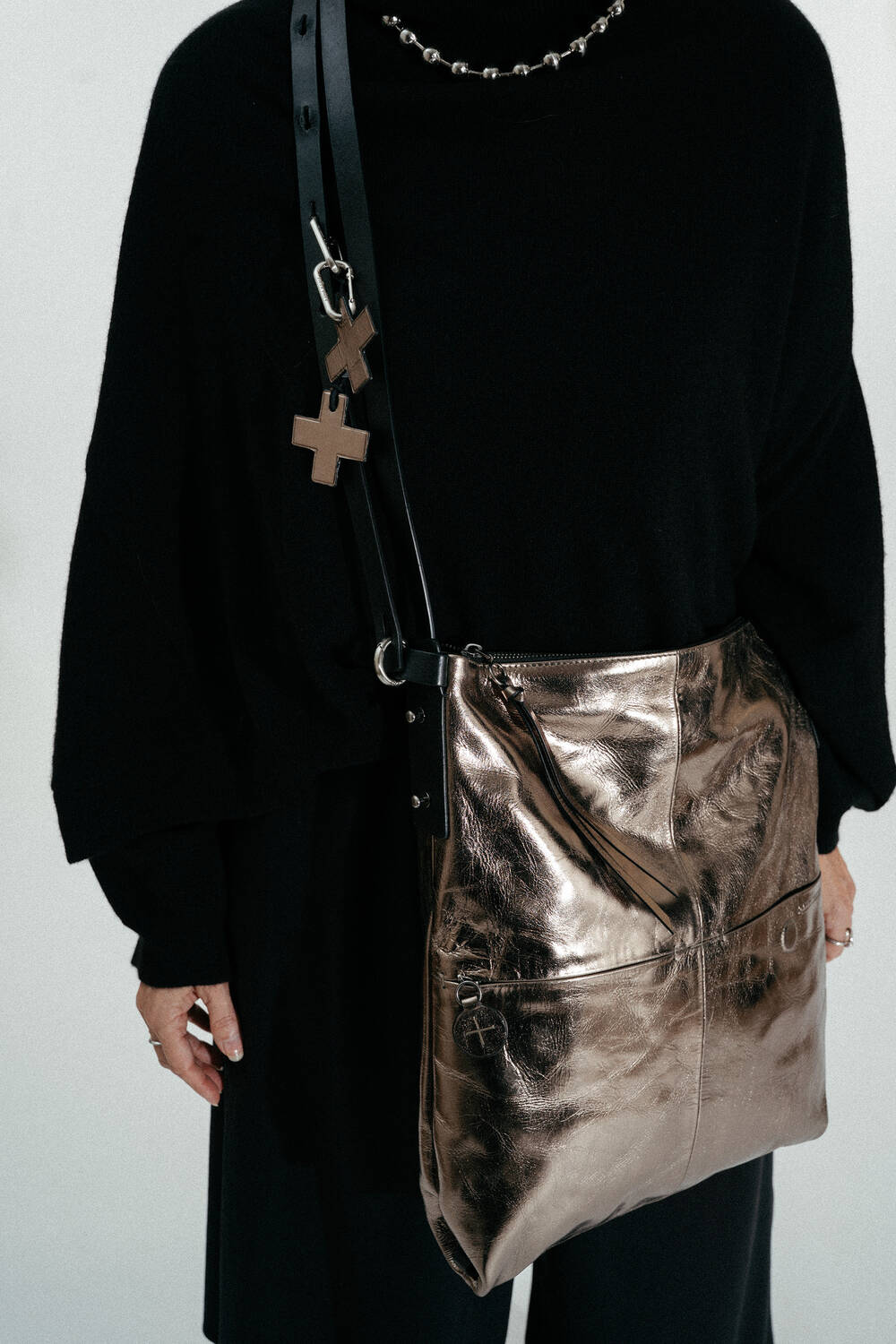 A person wearing a black outfit with an INA KENT metallic bronze shoulder bag featuring a black strap and cross-shaped charms.