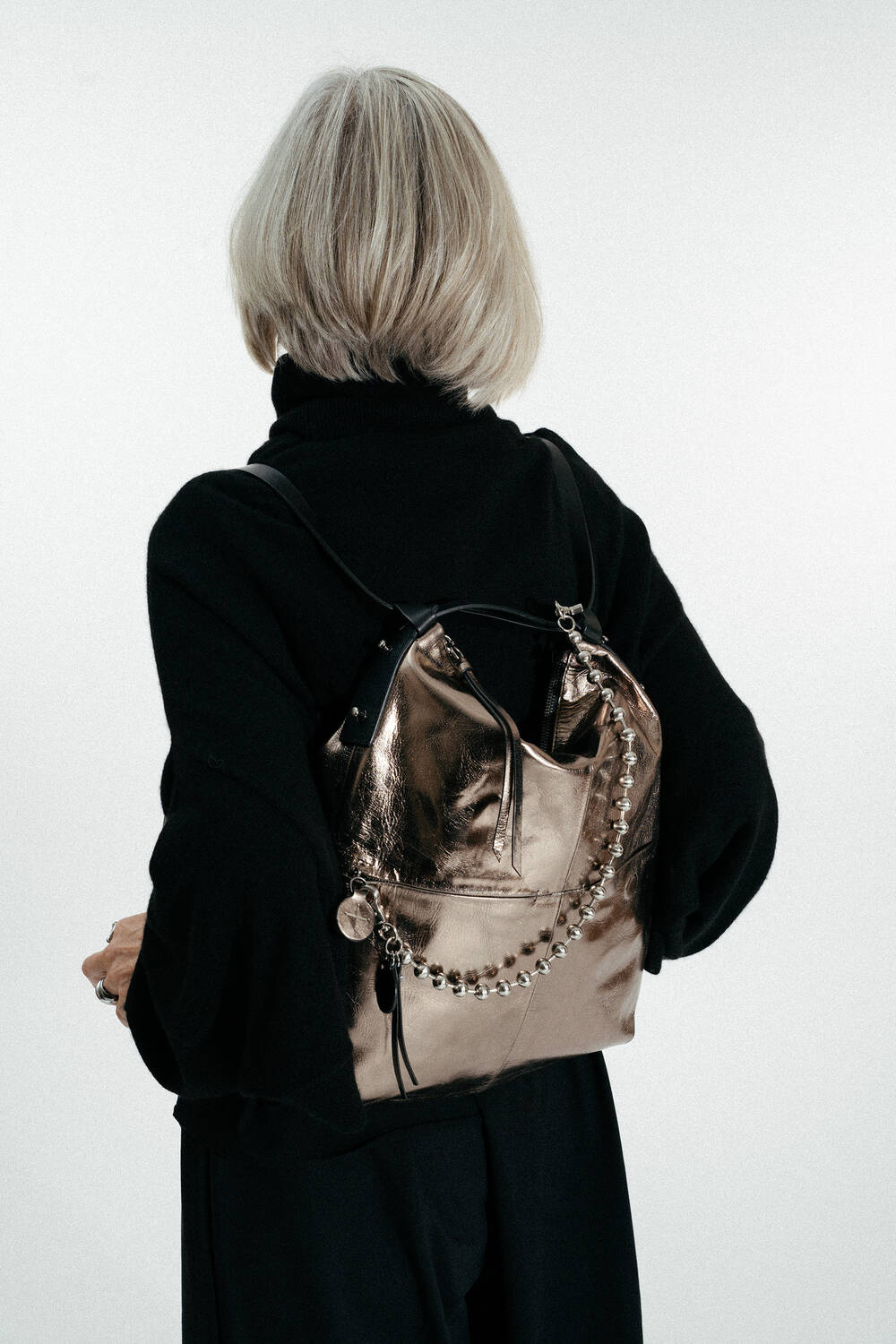 A person with short, gray hair wearing a black outfit faces away, showcasing an INA KENT metallic backpack with chain and charm details.