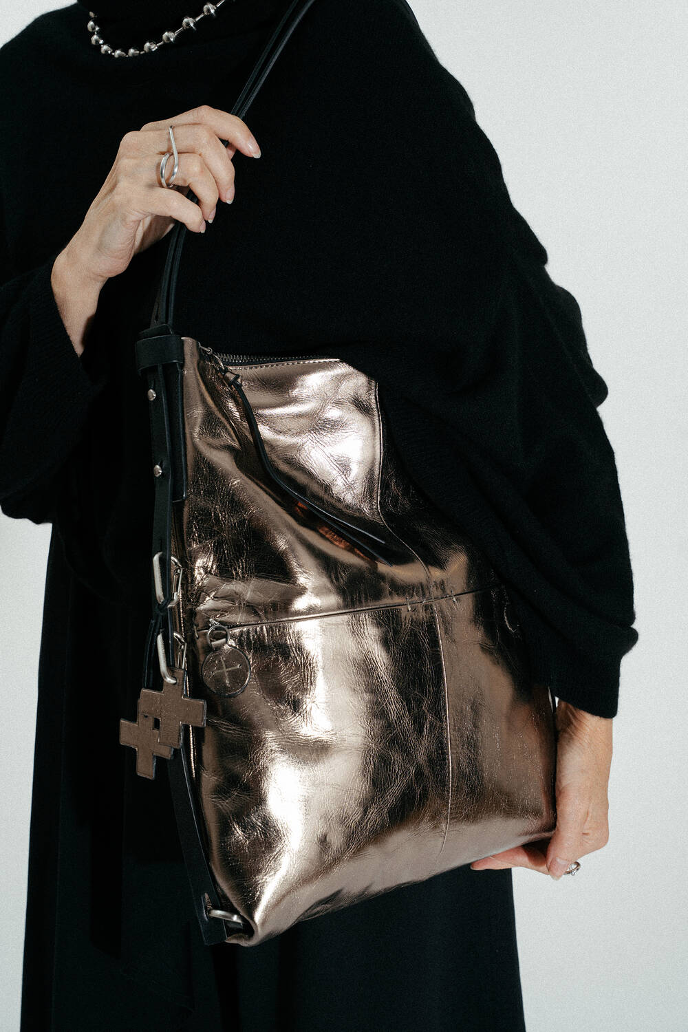 Person holding a large metallic INA KENT tote bag with a cross-shaped keychain, wearing a black outfit and a beaded necklace.