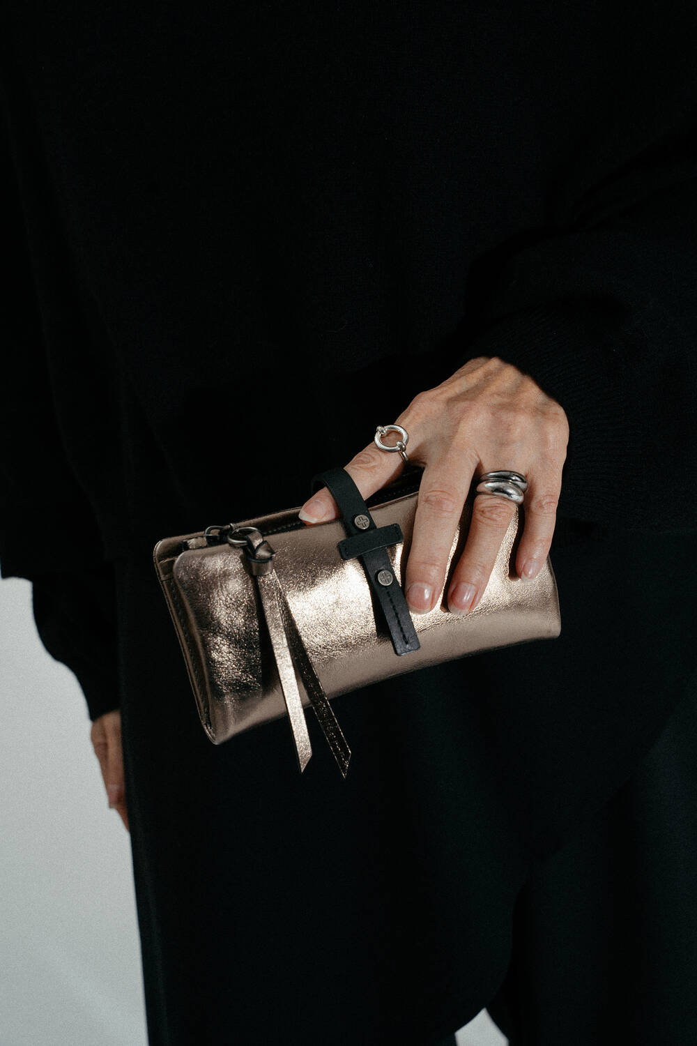 Eine Person, die silberne Ringe trägt, hält eine goldene INA KENT Metallic-Clutch-Tasche mit einem schwarzen Riemen und trägt schwarze Kleidung.