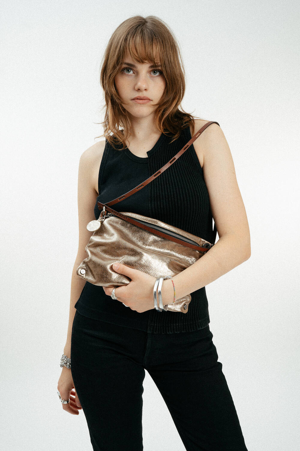 Eine Frau mit schulterlangen braunen Haaren trägt ein schwarzes ärmelloses Oberteil und eine schwarze Hose und hält eine goldene INA KENT-Metallic-Tasche mit einem braunen Riemen quer über ihren Körper.