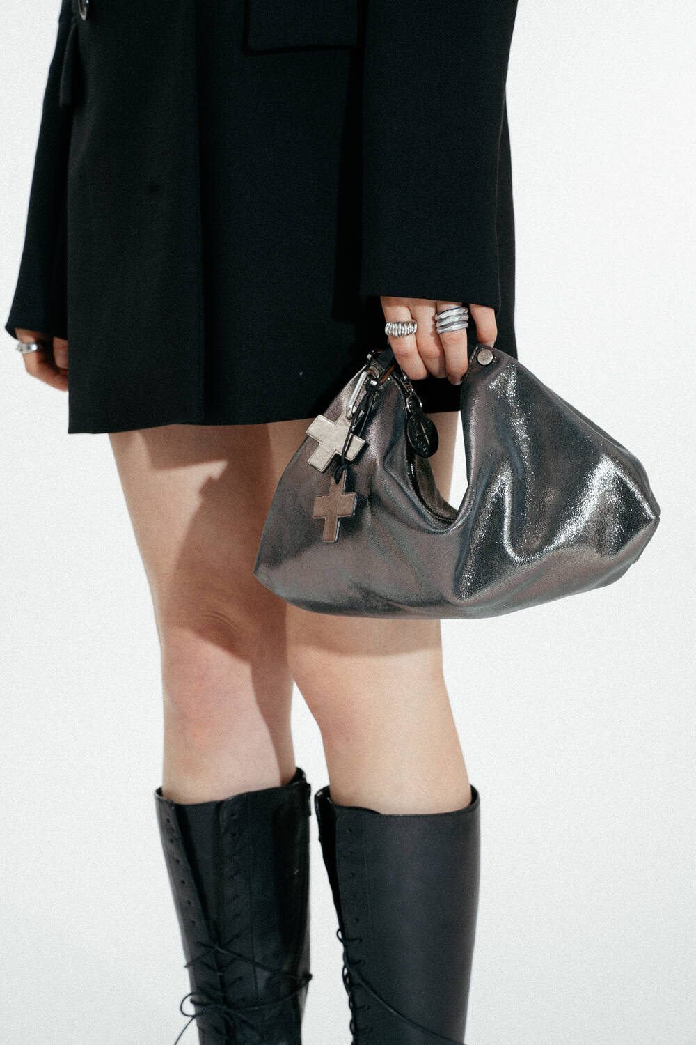 Eine Person, die einen schwarzen Blazer und schwarze kniehohe Stiefel trägt, hält eine silberglänzende INA KENT-Handtasche mit Schlüsselanhängern in der Hand.