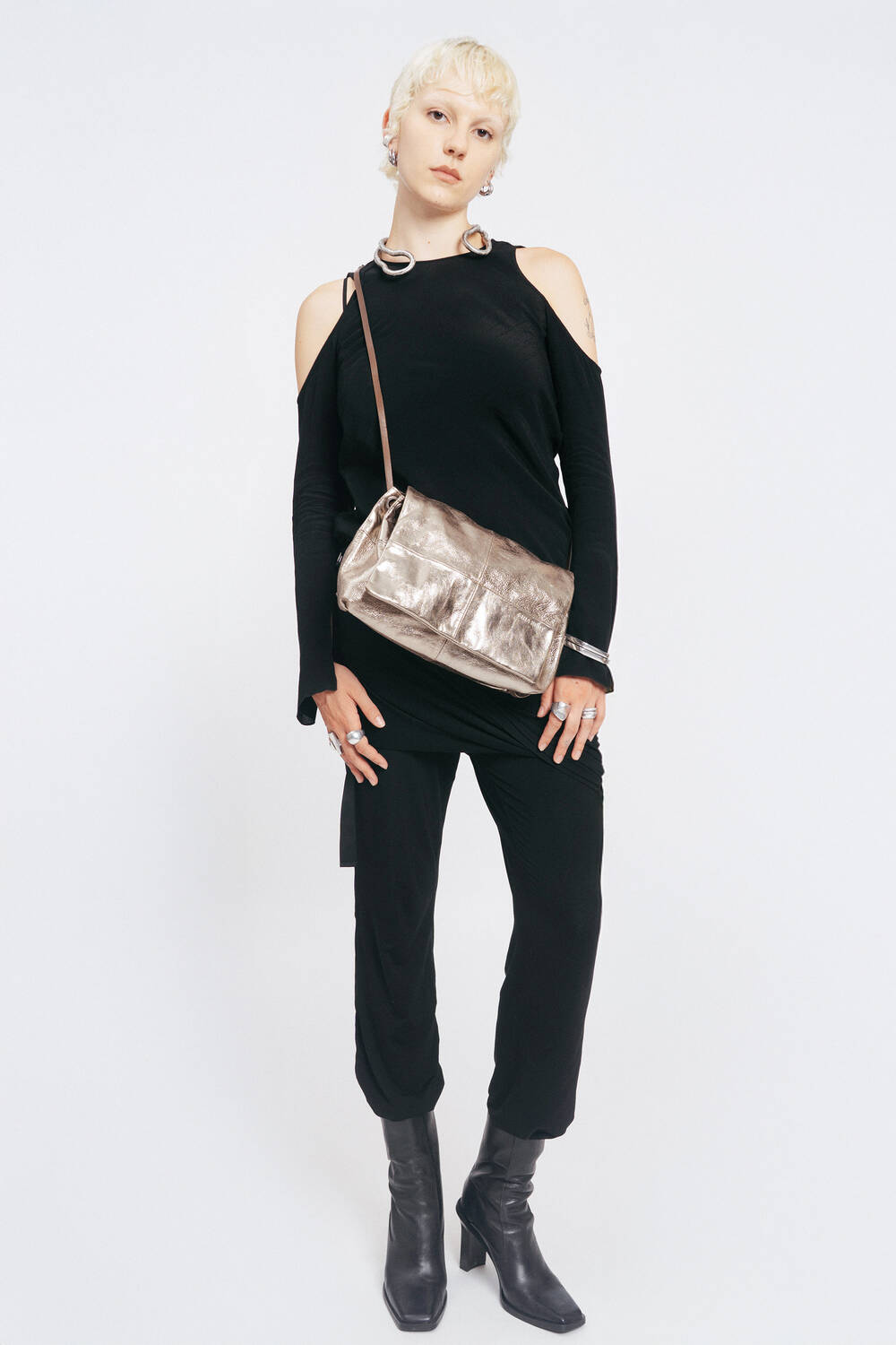 Person with short blond hair stands against a white background, wearing a black cold-shoulder outfit, black boots, and an INA KENT metallic crossbody bag.