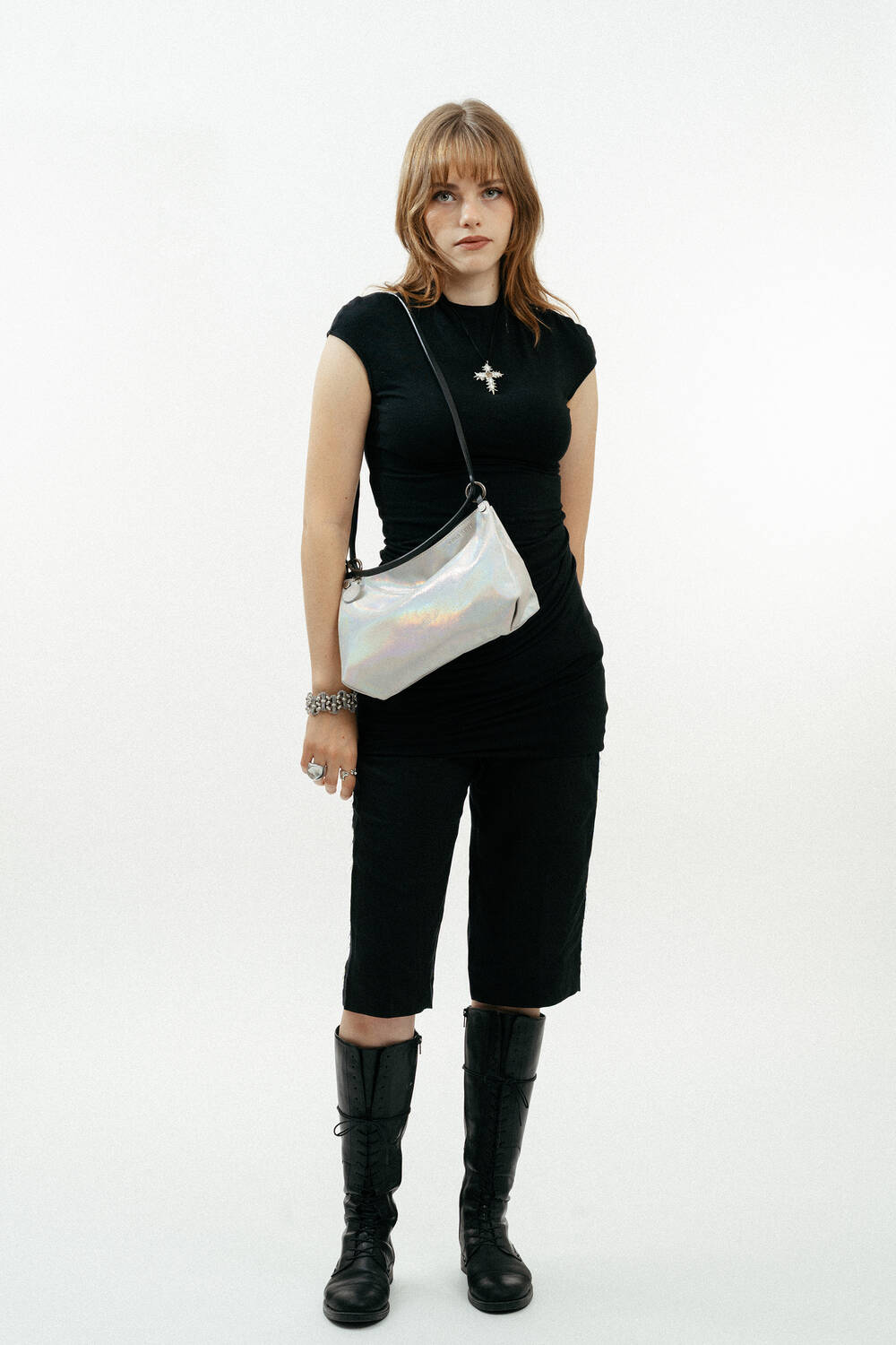 A person stands against a white background, wearing a black dress, black boots, a silver necklace, and carrying an INA KENT silver shoulder bag.