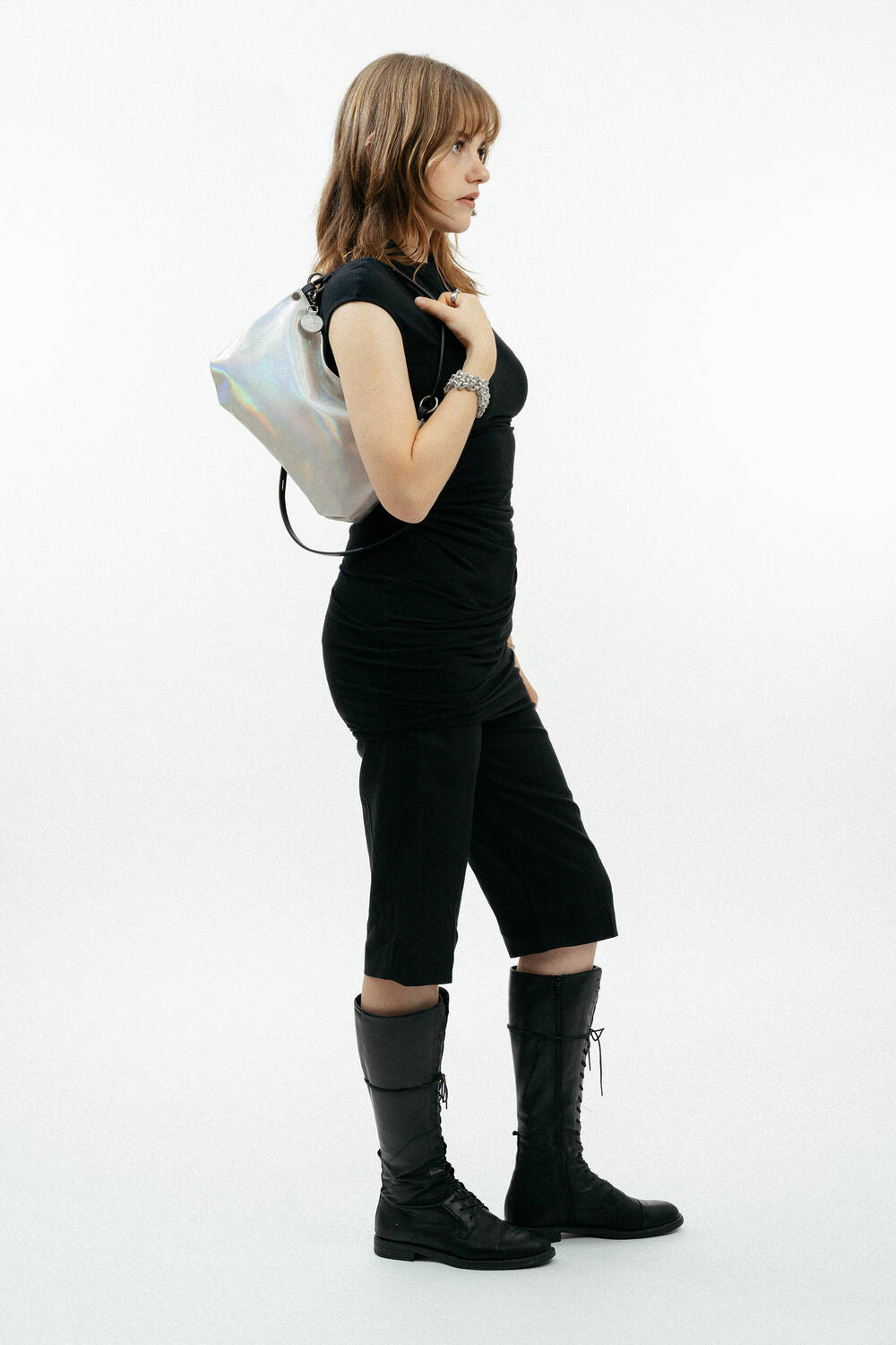 A woman stands in profile against a white background, wearing a black dress, knee-high black boots, and carrying an INA KENT silver shoulder bag.