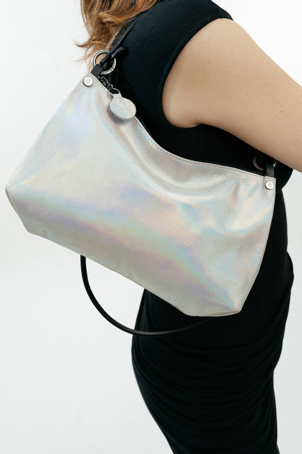 A person wearing a black sleeveless dress carries an INA KENT shiny, iridescent shoulder bag with a circular charm detail.