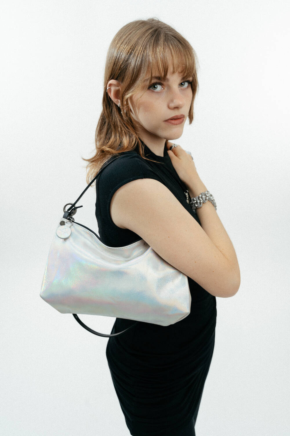 A woman in a black dress poses with an iridescent INA KENT shoulder bag against a plain white background.