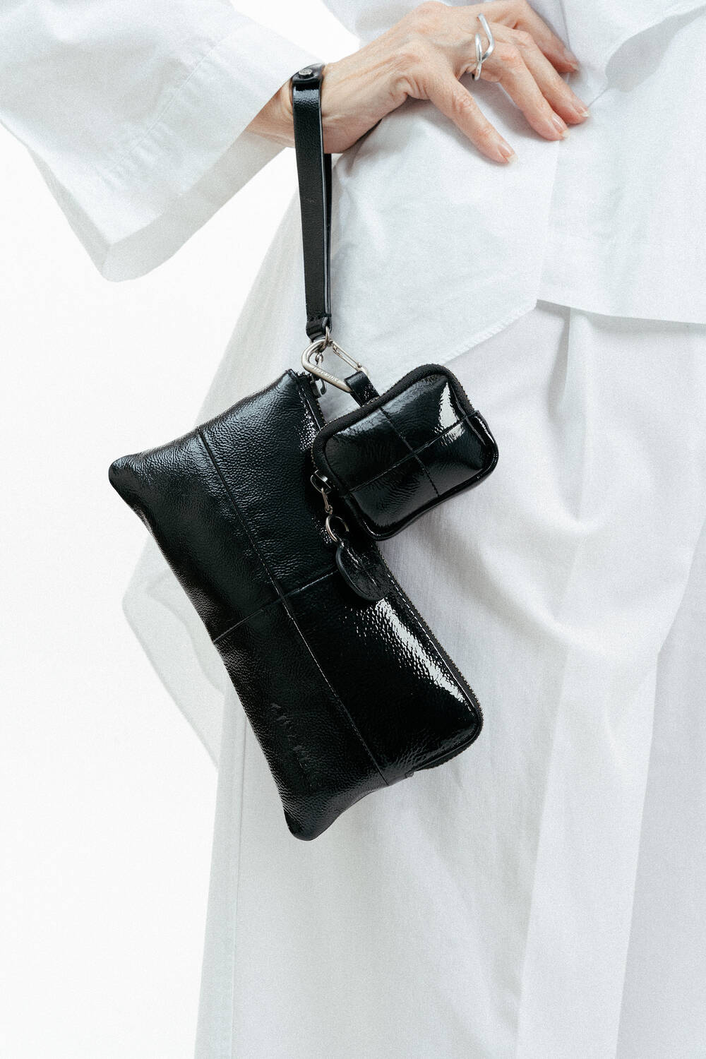 Eine weiß gekleidete Person hält eine glänzende, schwarze, rechteckige INA KENT-Clutch und ein dazu passendes kleines Täschchen, die beide an einem Armband befestigt sind.