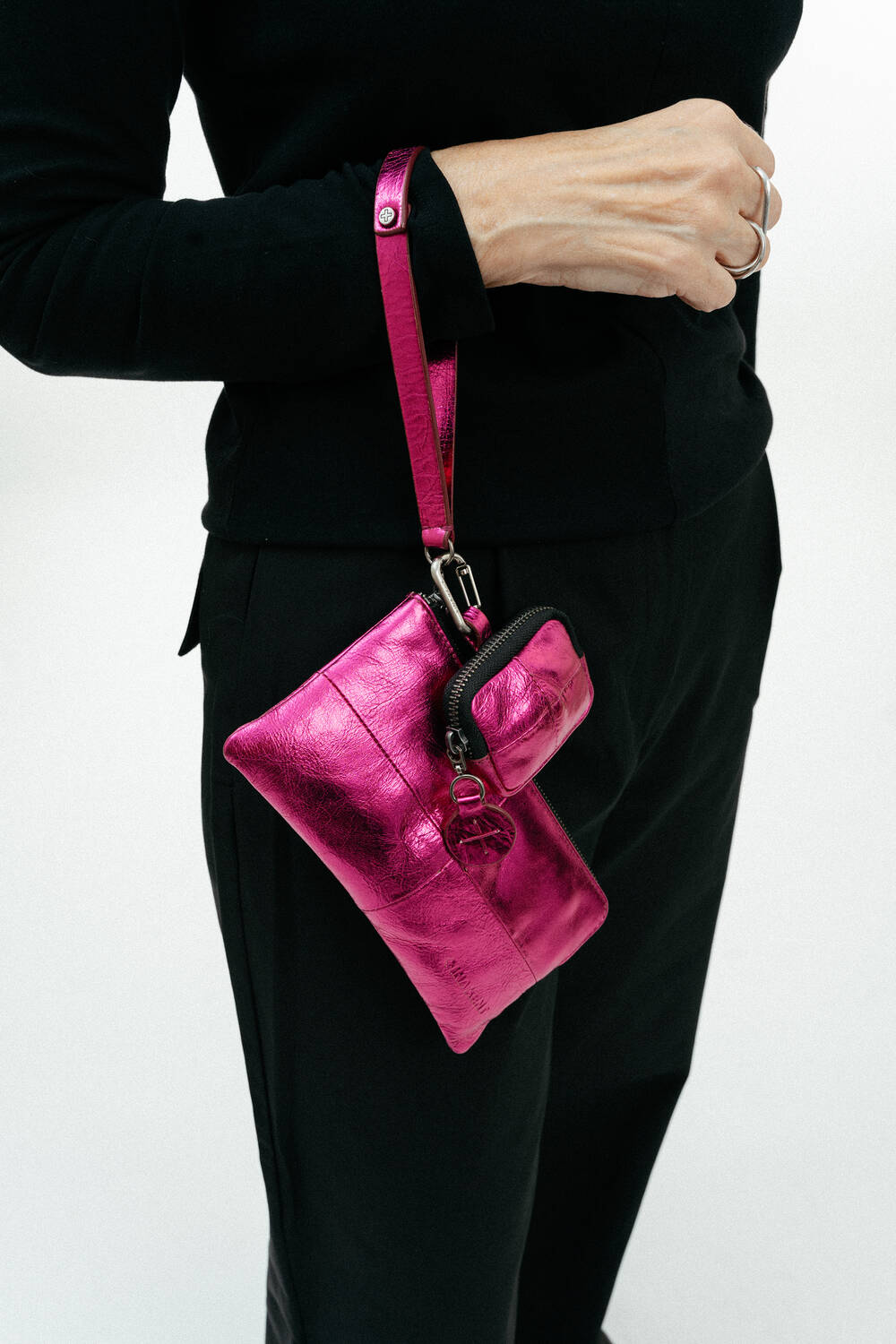 Eine schwarz gekleidete Person hält eine metallisch pinkfarbene INA KENT-Armbandtasche mit einem dazu passenden kleinen Beutel vor einem schlichten weißen Hintergrund.