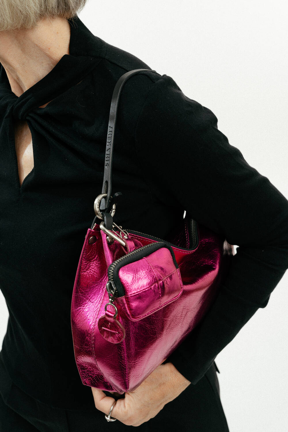 Eine schwarz gekleidete Person hält eine rosafarbene Metallic-Handtasche von INA KENT mit einer kleinen passenden Tasche und einem schwarzen Schulterriemen.