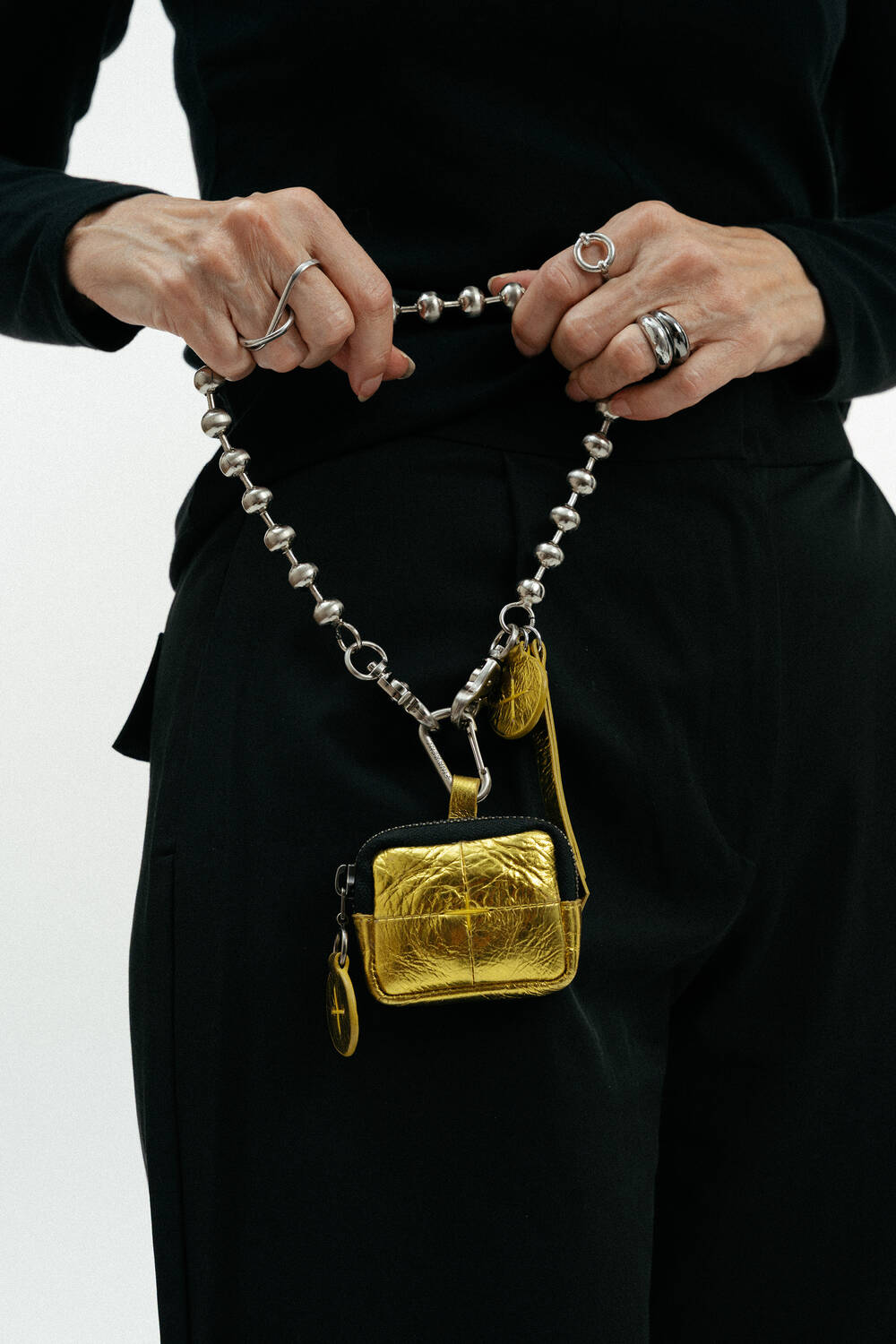 Person wearing a black outfit holds a beaded chain with an INA KENT metallic gold pouch and keychain attached, showcasing multiple silver rings on their fingers.