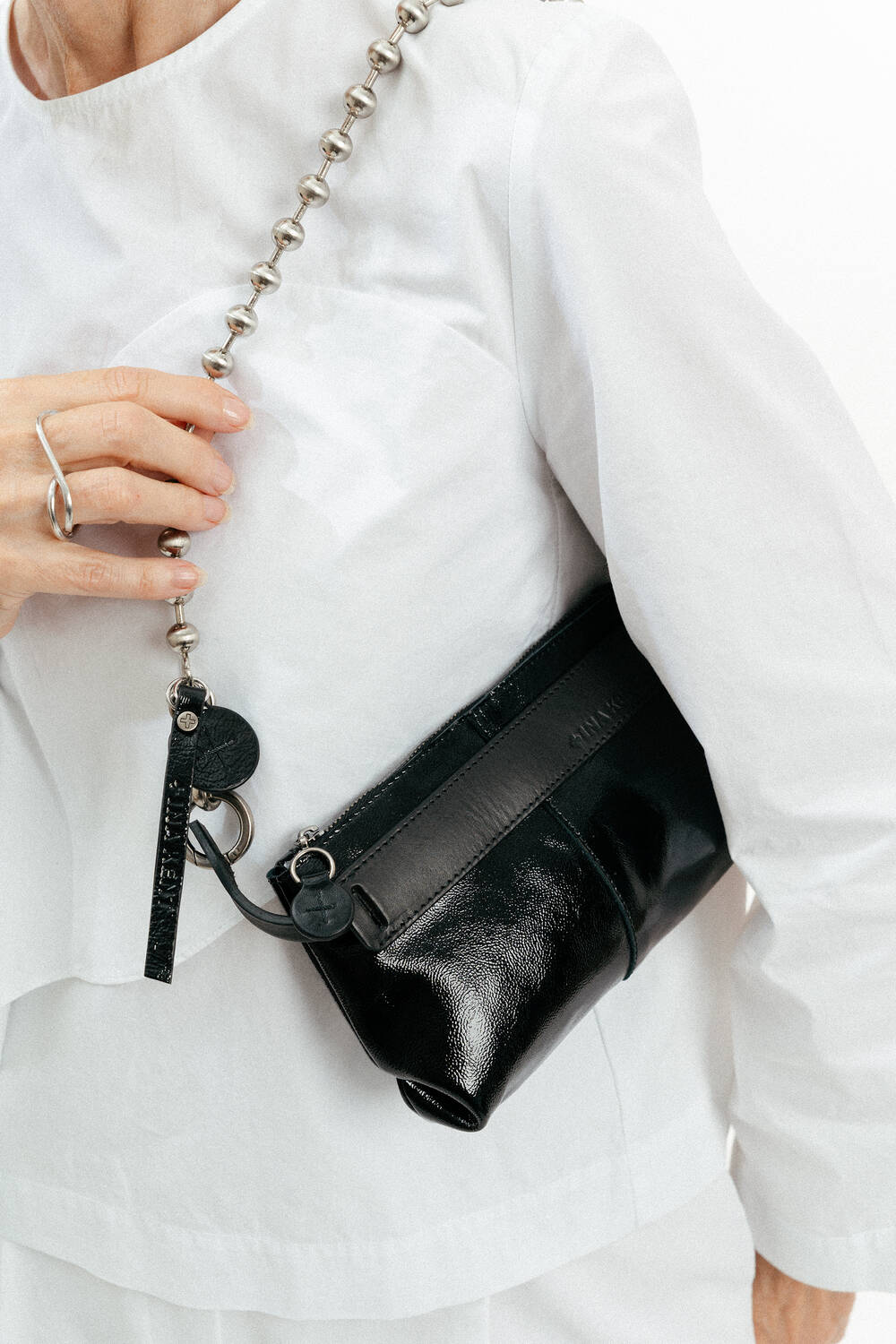 Person wearing a white outfit holds an INA KENT black leather shoulder bag with a silver chain strap and a keychain accessory.