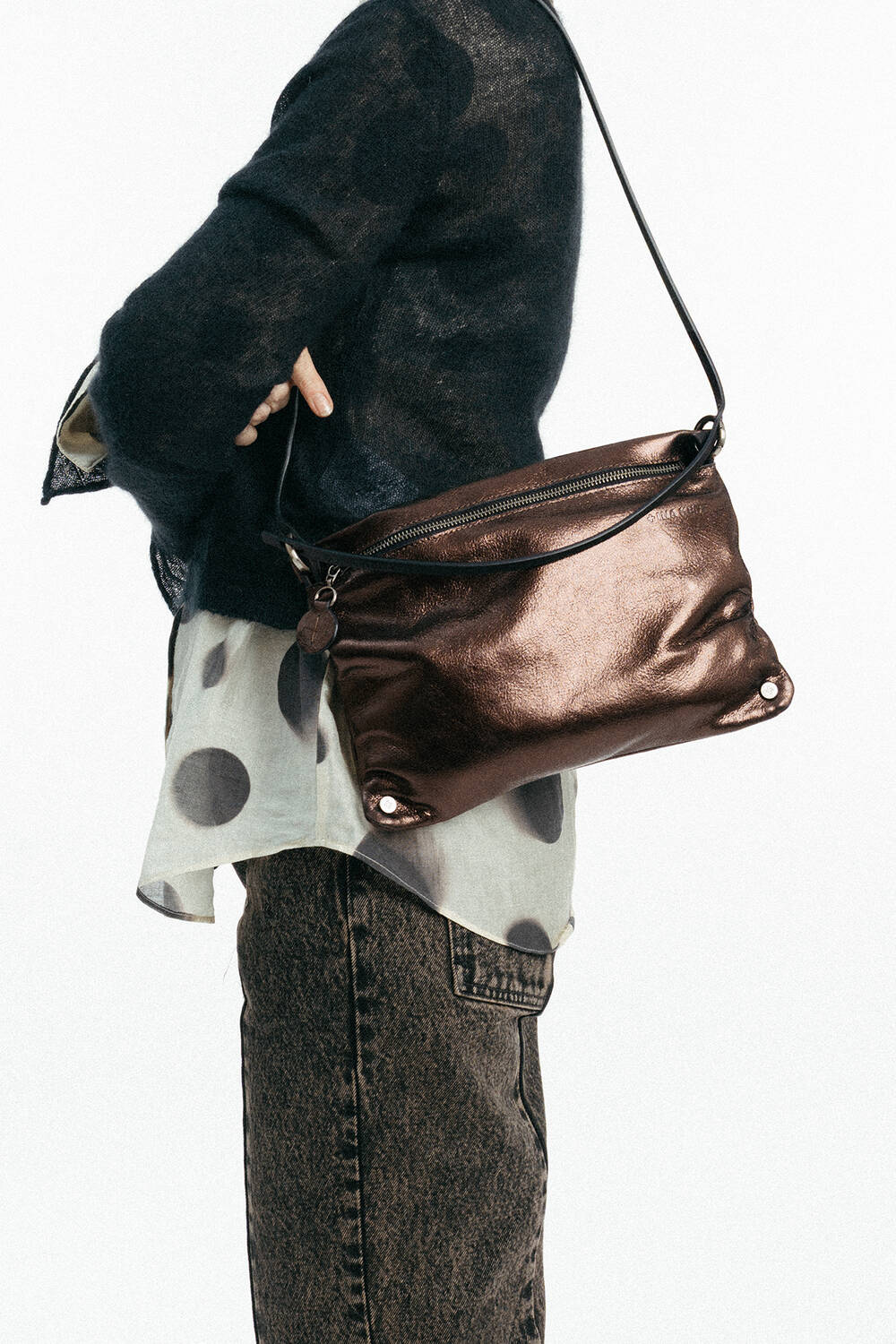 Person, die einen durchsichtigen schwarzen Pullover über einem gepunkteten Hemd trägt und eine INA KENT Metallic-Bronze-Umhängetasche in der Hand hält; dunkle Jeans sind zu sehen.