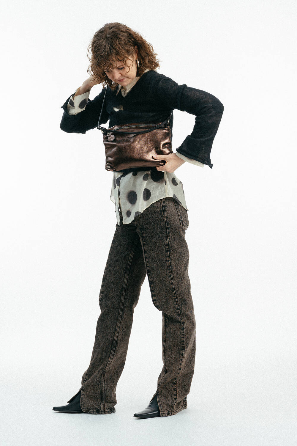 Person mit lockigem Haar, die eine Brille, eine schwarze Jacke, ein gepunktetes Hemd, braune Jeans und schwarze Schuhe trägt, hält eine INA KENT Metallic-Umhängetasche. Weißer Hintergrund.