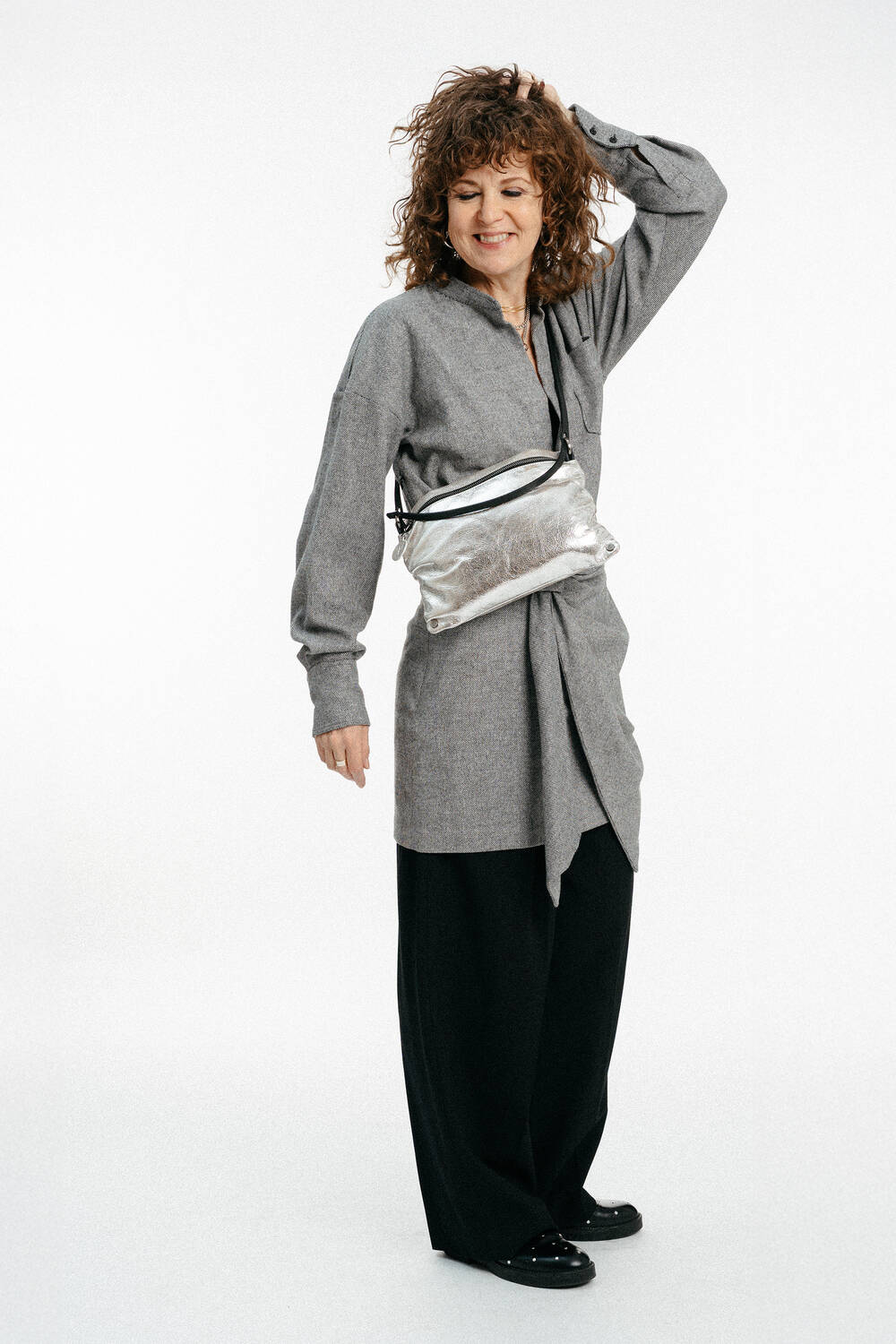 Eine Frau mit lockigem Haar, die ein graues langes Hemd, eine schwarze Hose mit weitem Bein, schwarze Schuhe und eine INA KENT Metallic-Crossbody-Tasche trägt, steht vor einem weißen Hintergrund und lächelt mit einer Hand in ihrem Haar.